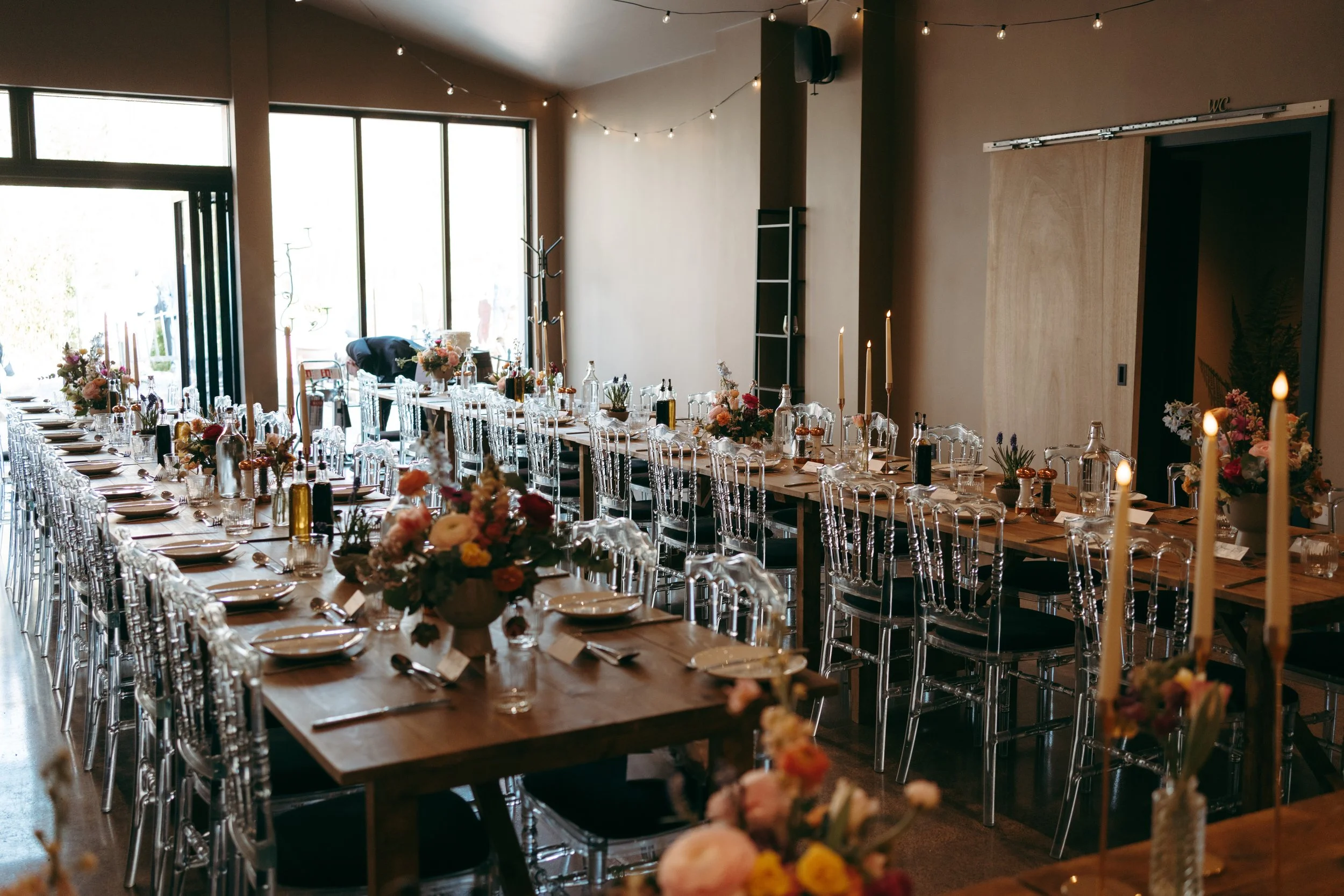 A decorated event reception hall with long wooden tables, clear acrylic chairs, floral centerpieces, tall candles, and string lights, with large windows letting in natural light.