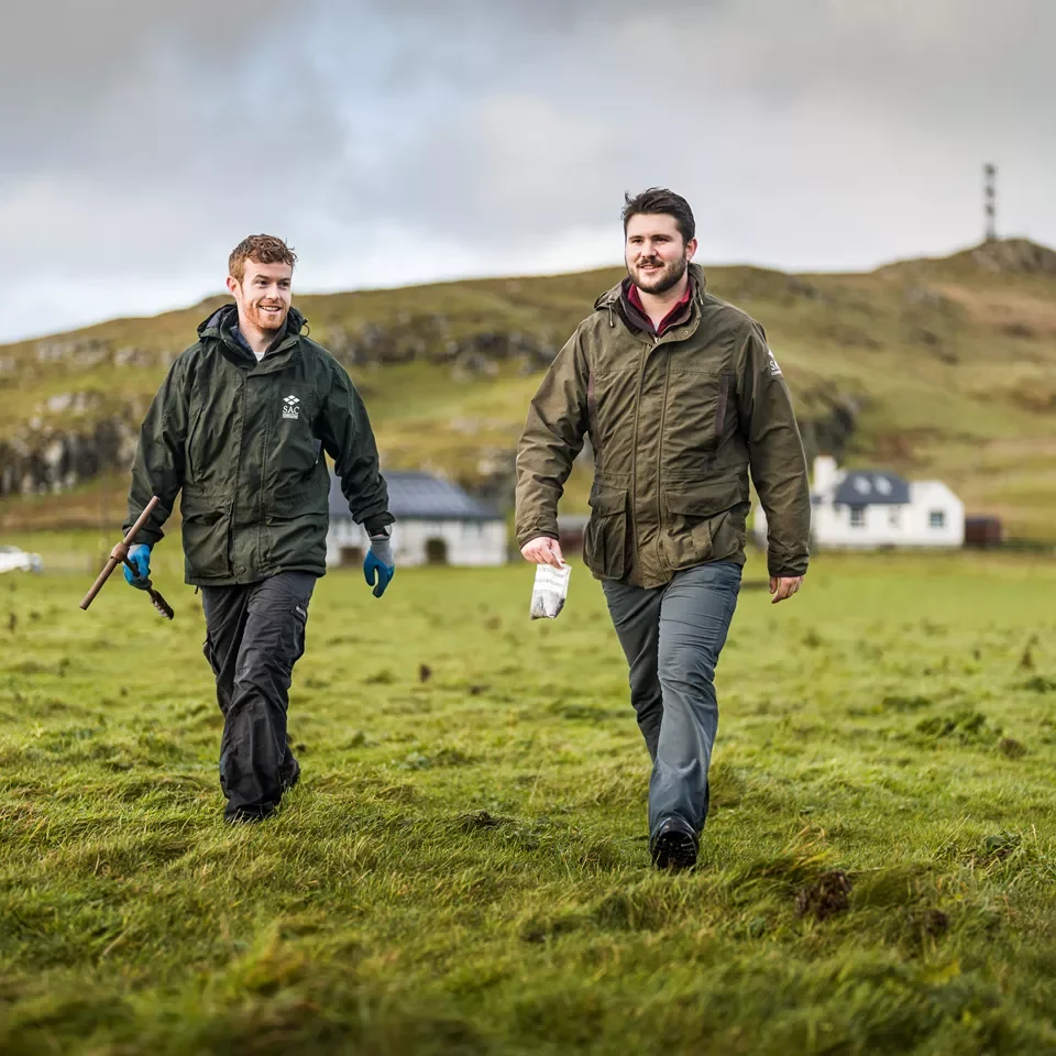 Two men walking on a grassy field in a rural area with hills and houses in the background. They are dressed in outdoor jackets and appear to be enjoying a hike or walk.