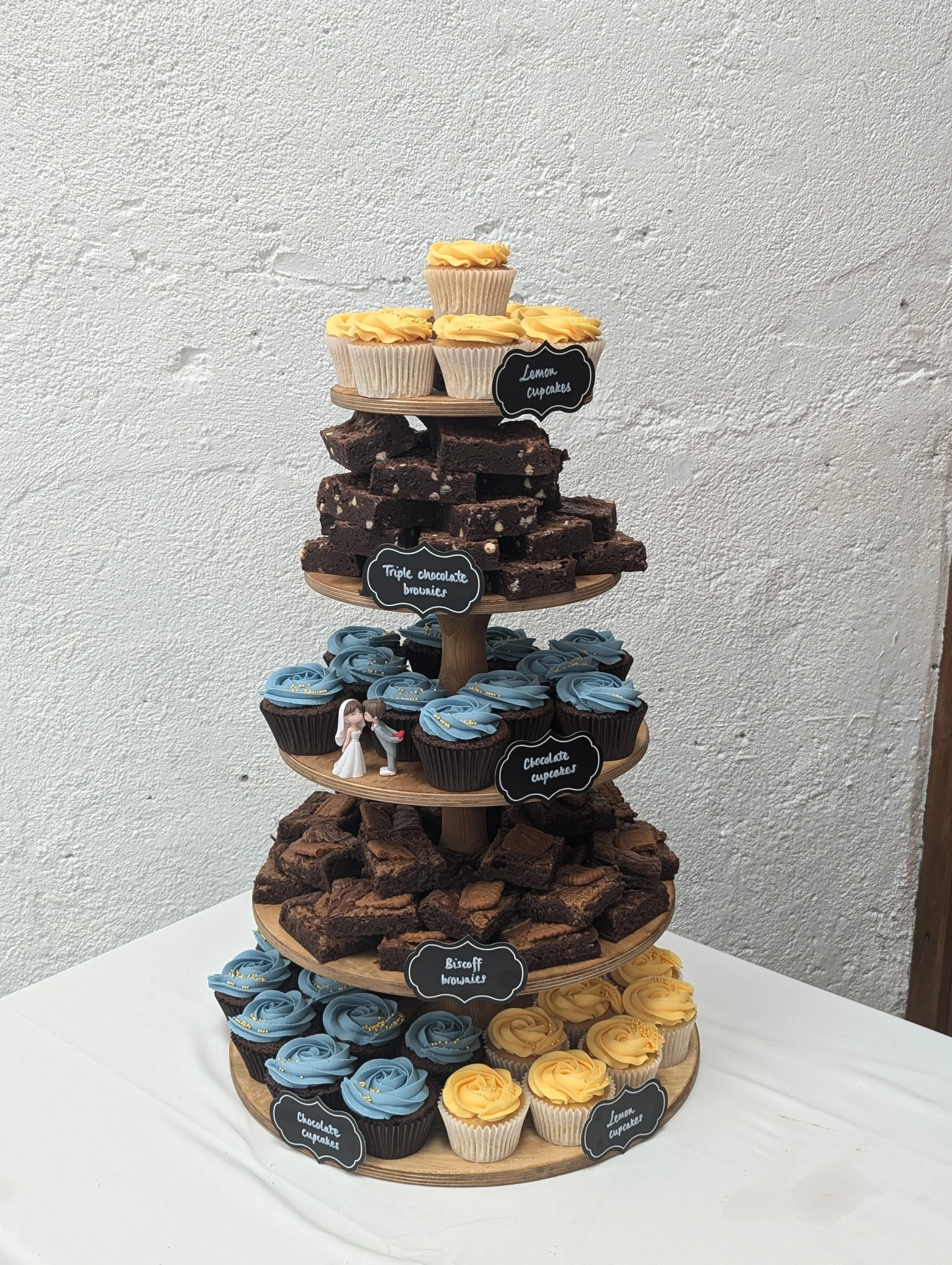 Multi-tiered dessert display with cupcakes, brownies, and cookies on a white table against a textured white wall.