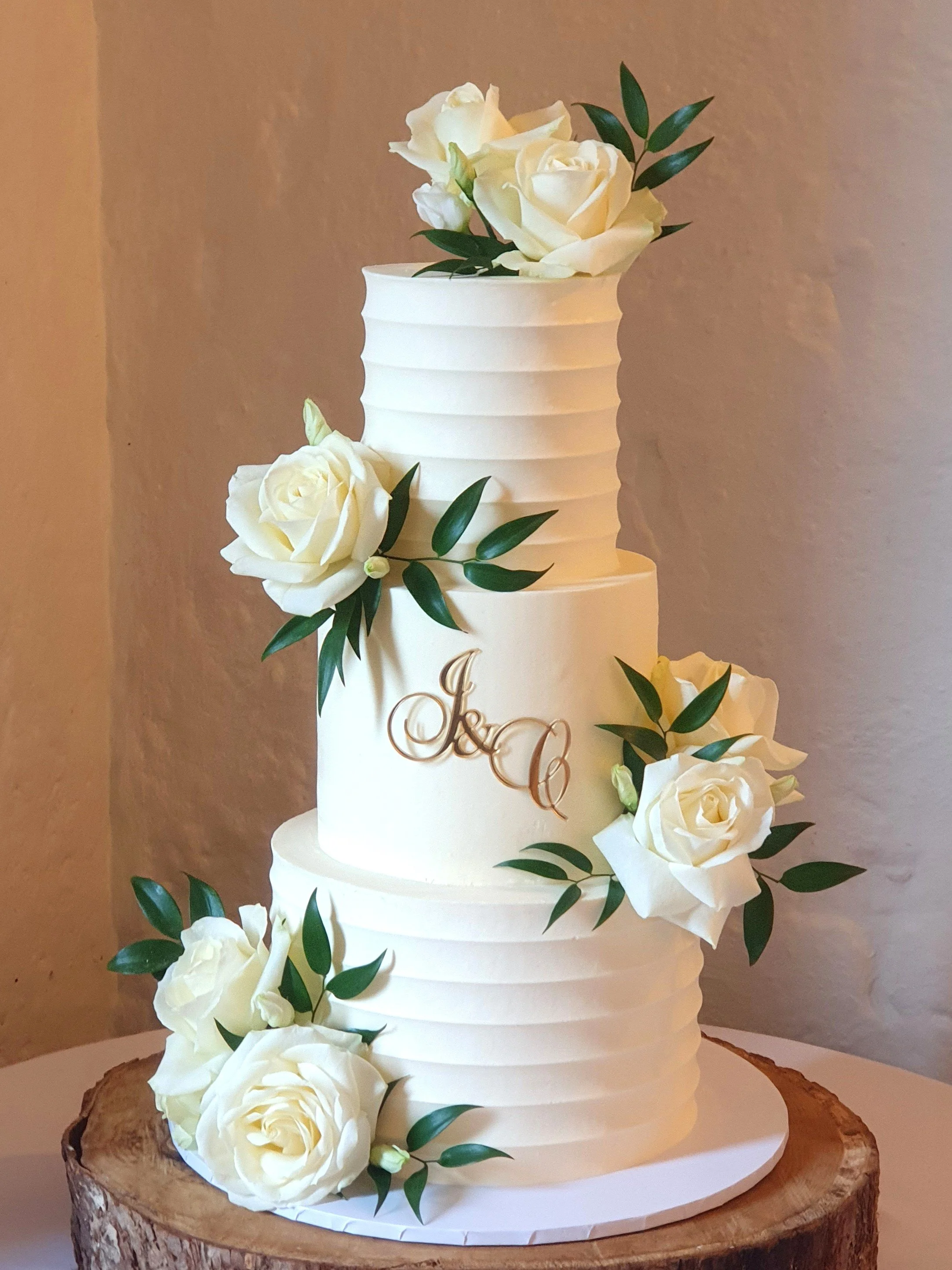 A white, three-tiered wedding cake at Priston Mill, decorated with white roses and green leaves, placed on a log slice.