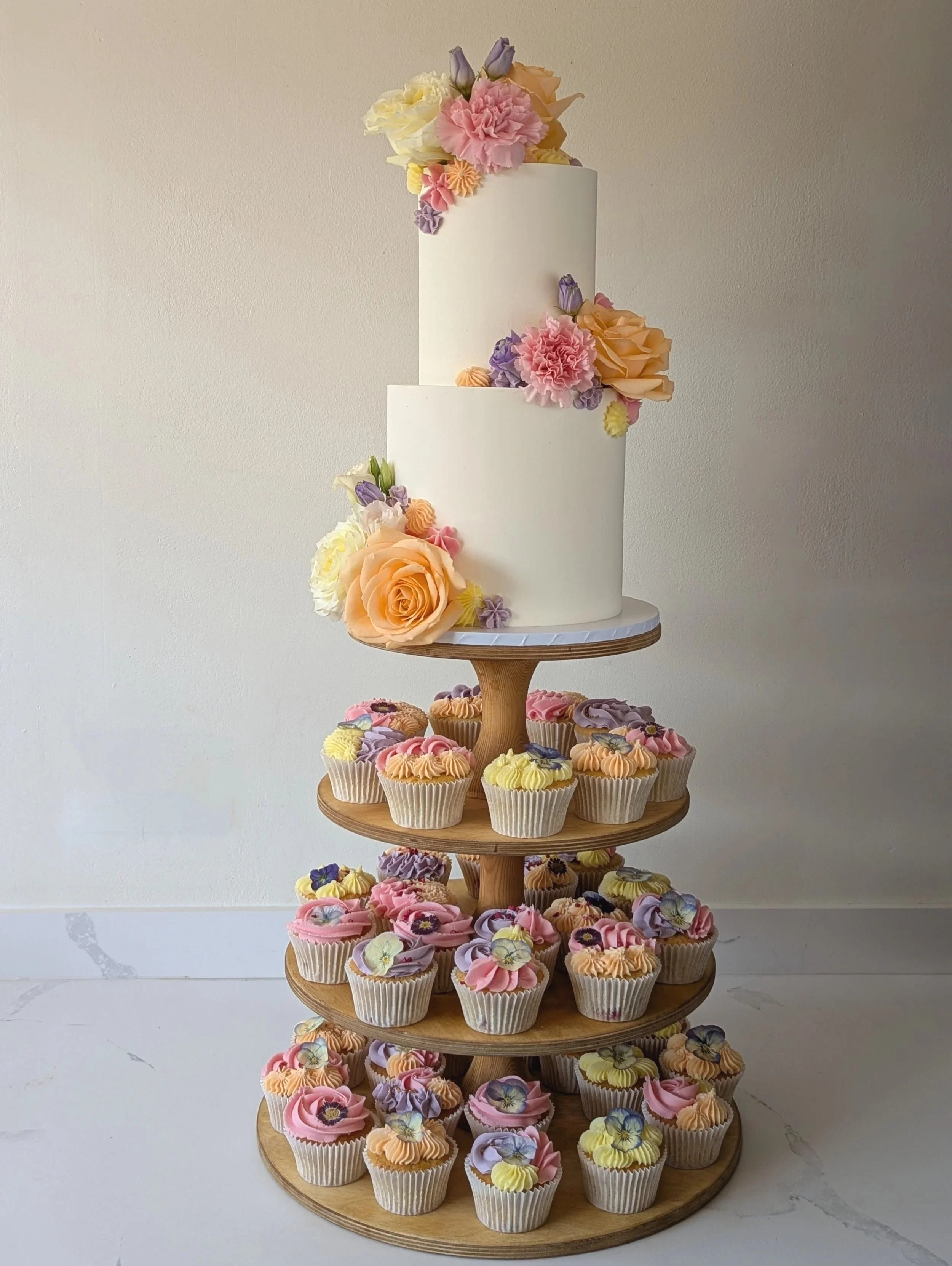A multi-tiered cake decorated with pastel-colored flowers, surrounded by cupcakes on a wooden stand. Syrencot, Wiltshire. 