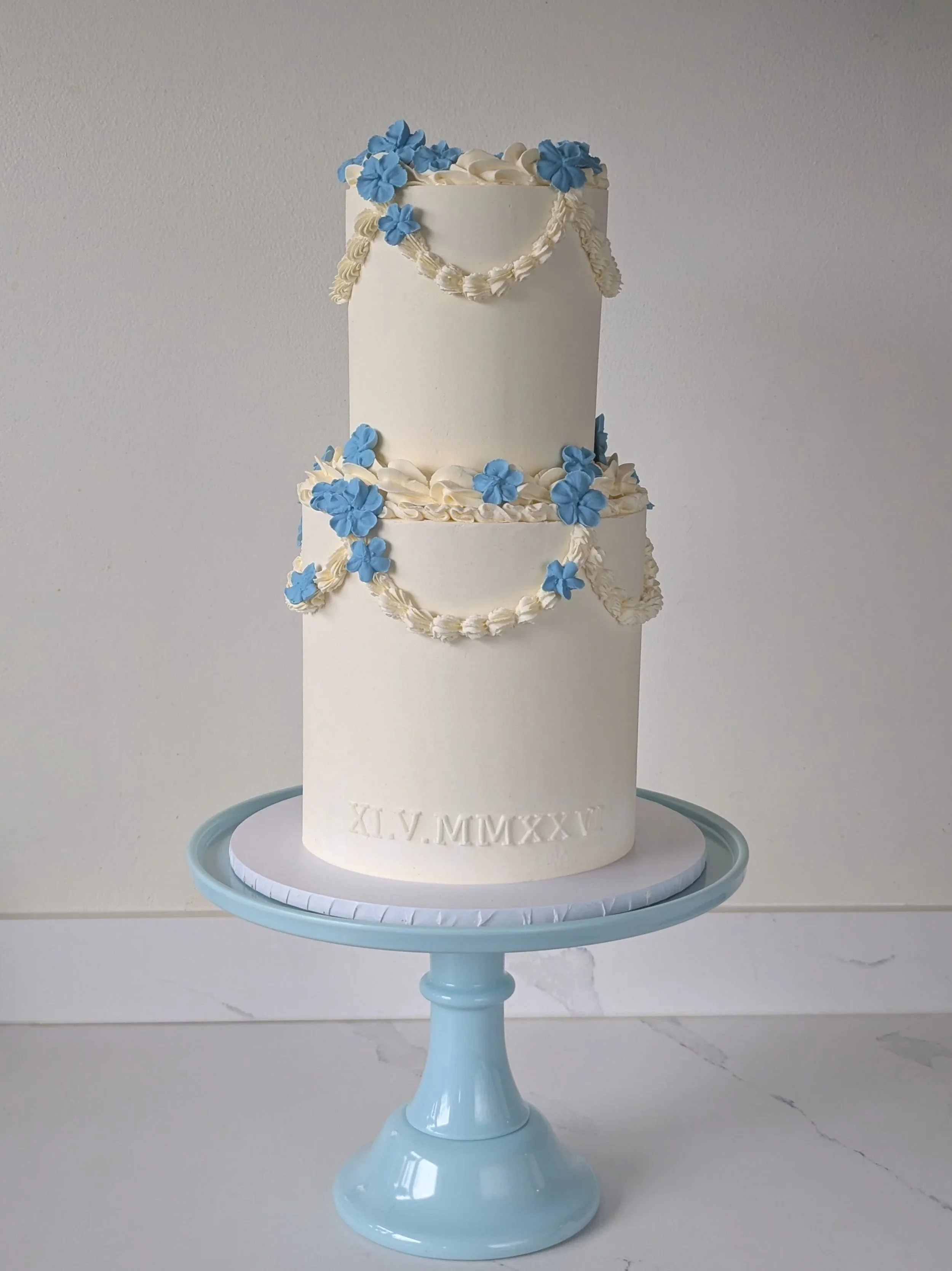 A two-tiered white cake decorated with blue flowers and cream-colored piping, placed on a light blue cake stand against a neutral background.