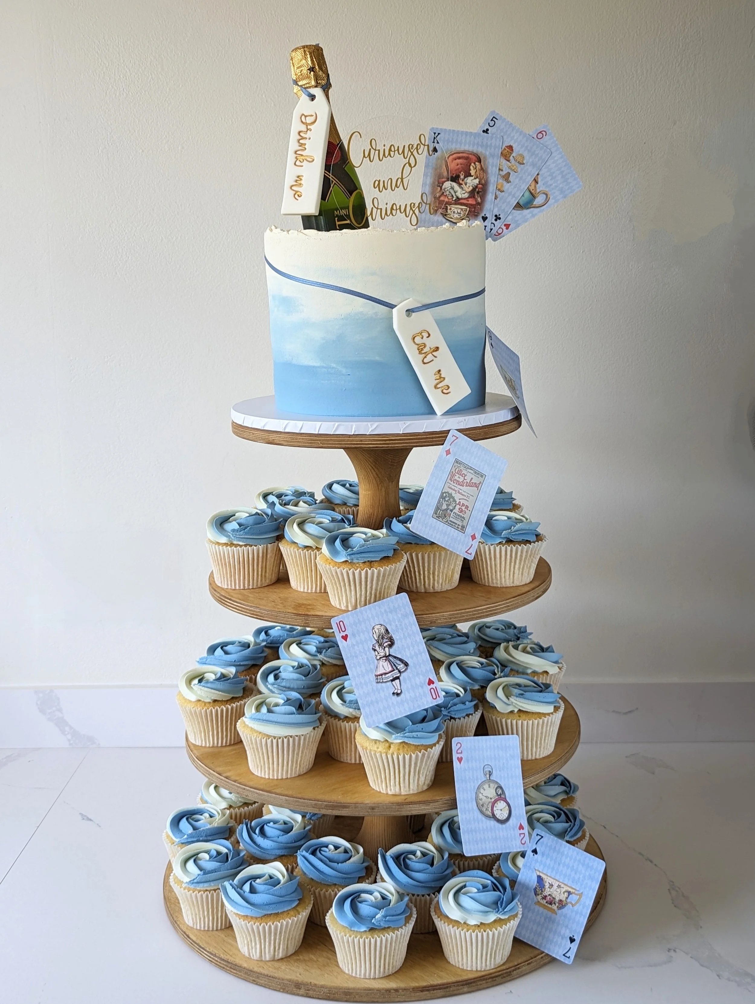A multi-tiered dessert display shaped like the storybook scene from Alice in Wonderland, featuring cupcakes with blue and white swirl frosting, a cake with painted sky and clouds, and playing cards with Alice in Wonderland illustrations and character