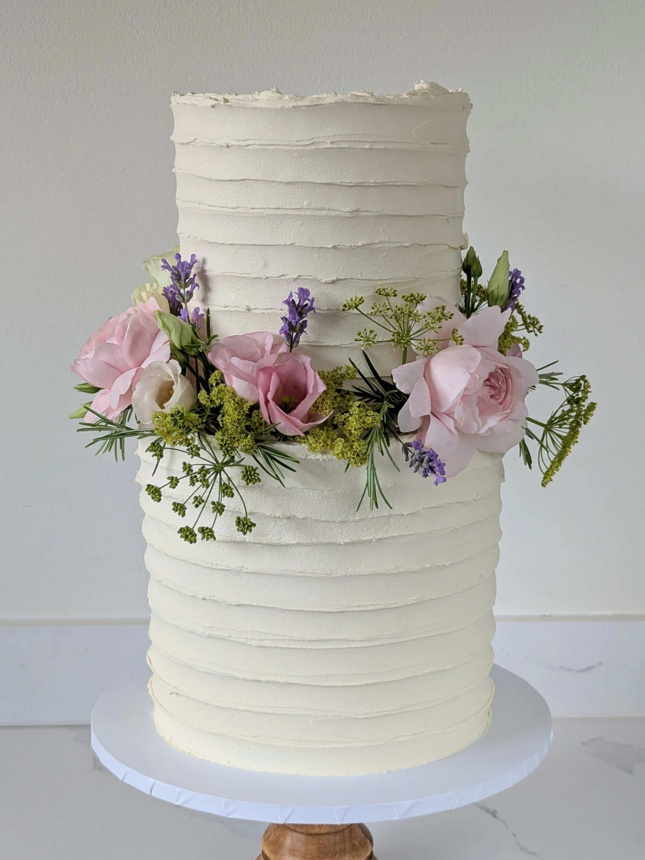 A tall, white, textured cake with horizontal ridges, decorated with pink and purple flowers and green foliage, placed on a round white cake board, on a cake stand.