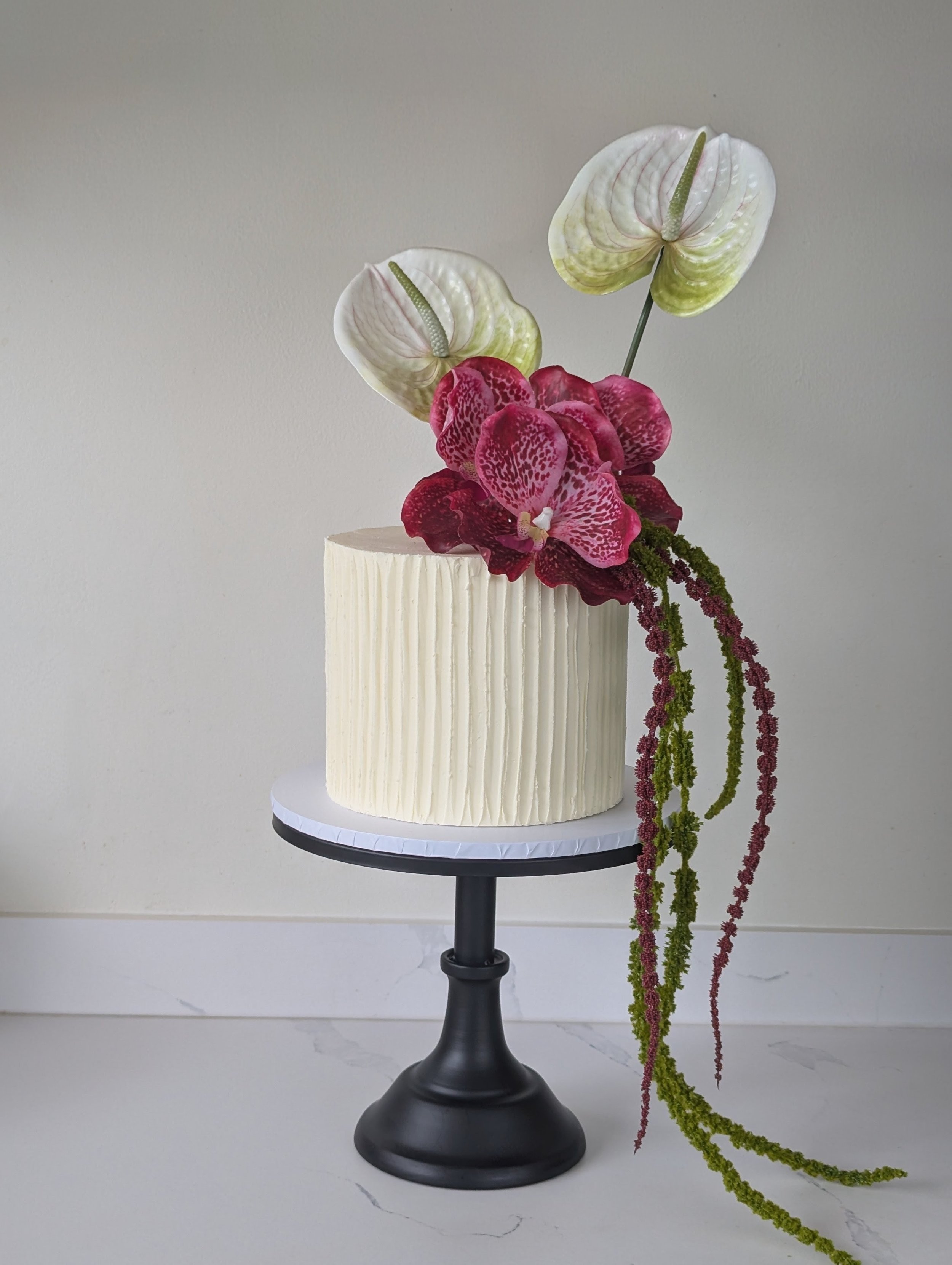 A cake decorated with an arrangement of orchids, featuring white anthuriums and pink and red spotted orchids with long trailing green and pink amaranthus, placed on a black pedestal stand.