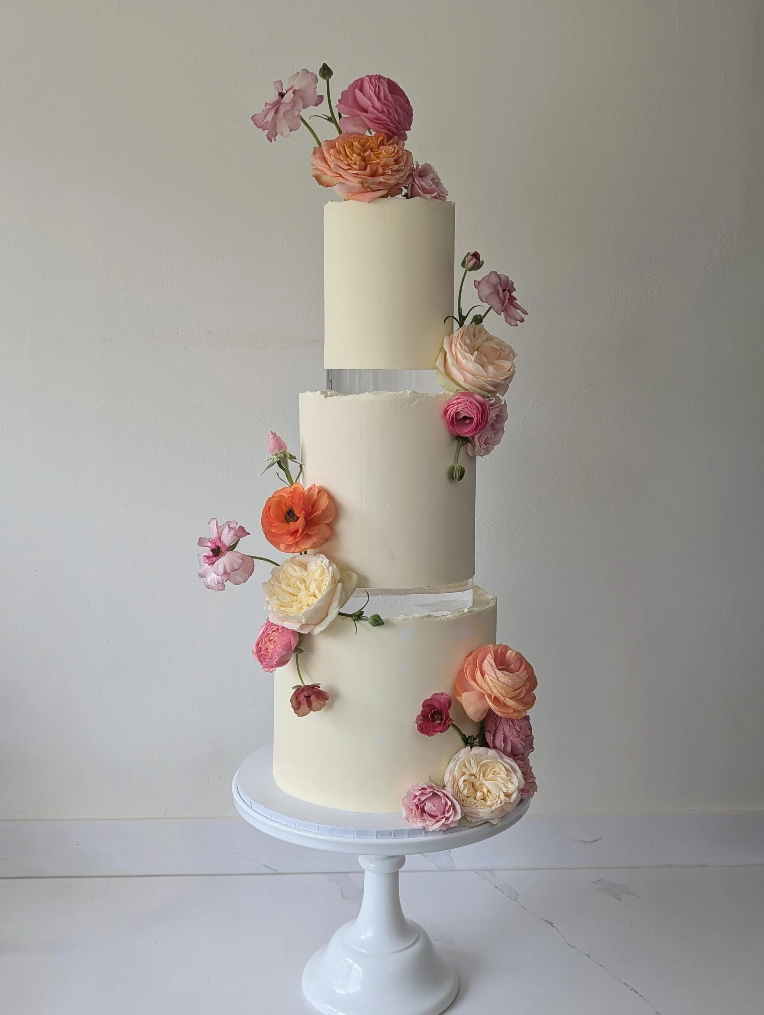 A three-tiered white wedding cake decorated with pink, peach, and cream-colored flowers, with some flowers cascading down the side. The cake is on a white pedestal cake stand against a plain light gray background.