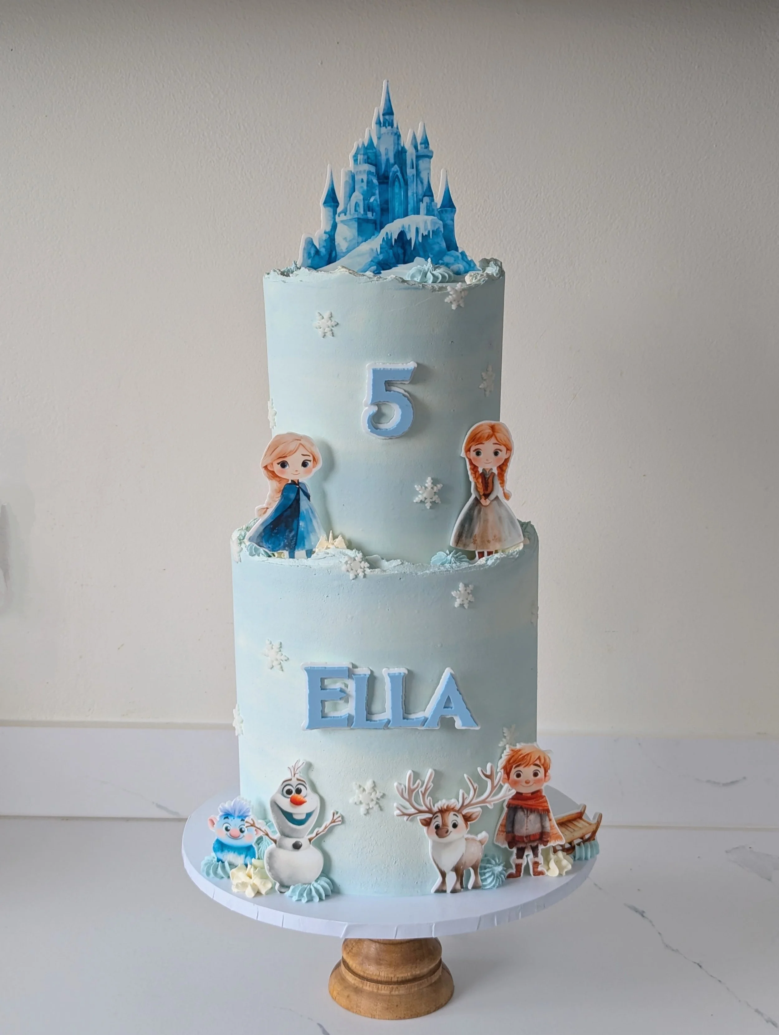 A three-tiered birthday cake decorated with 'Frozen' characters, snowflakes, and a castle topper. The cake has the name 'ELLA' on it, with a 5th birthday theme.