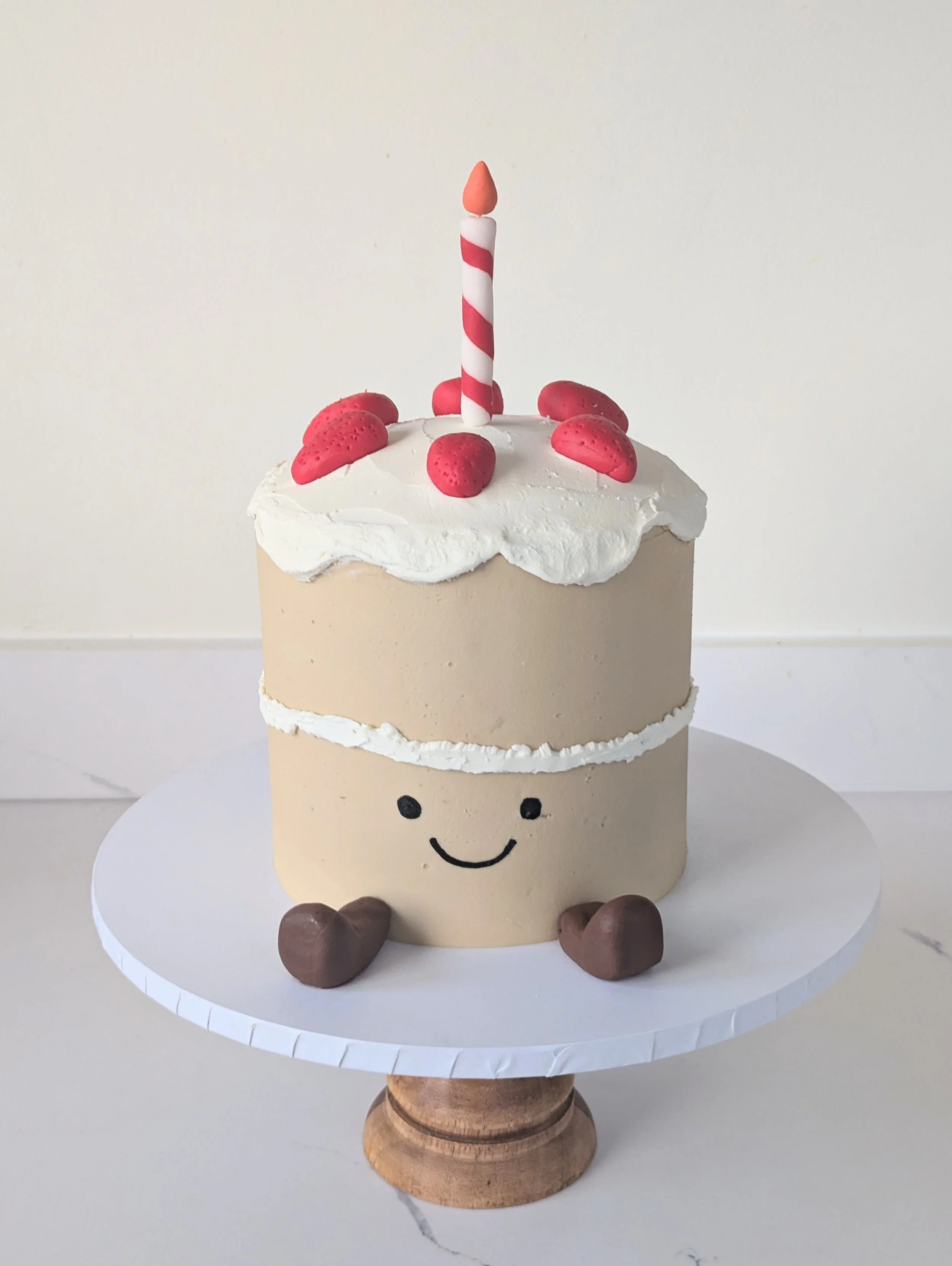 A cake decorated like a smiling face with a white top layer, a beige middle layer, and a cream-colored bottom layer, sitting on a white cake stand. The cake has a face with black dot eyes and a curved line smile. It is topped with a birthday candle a