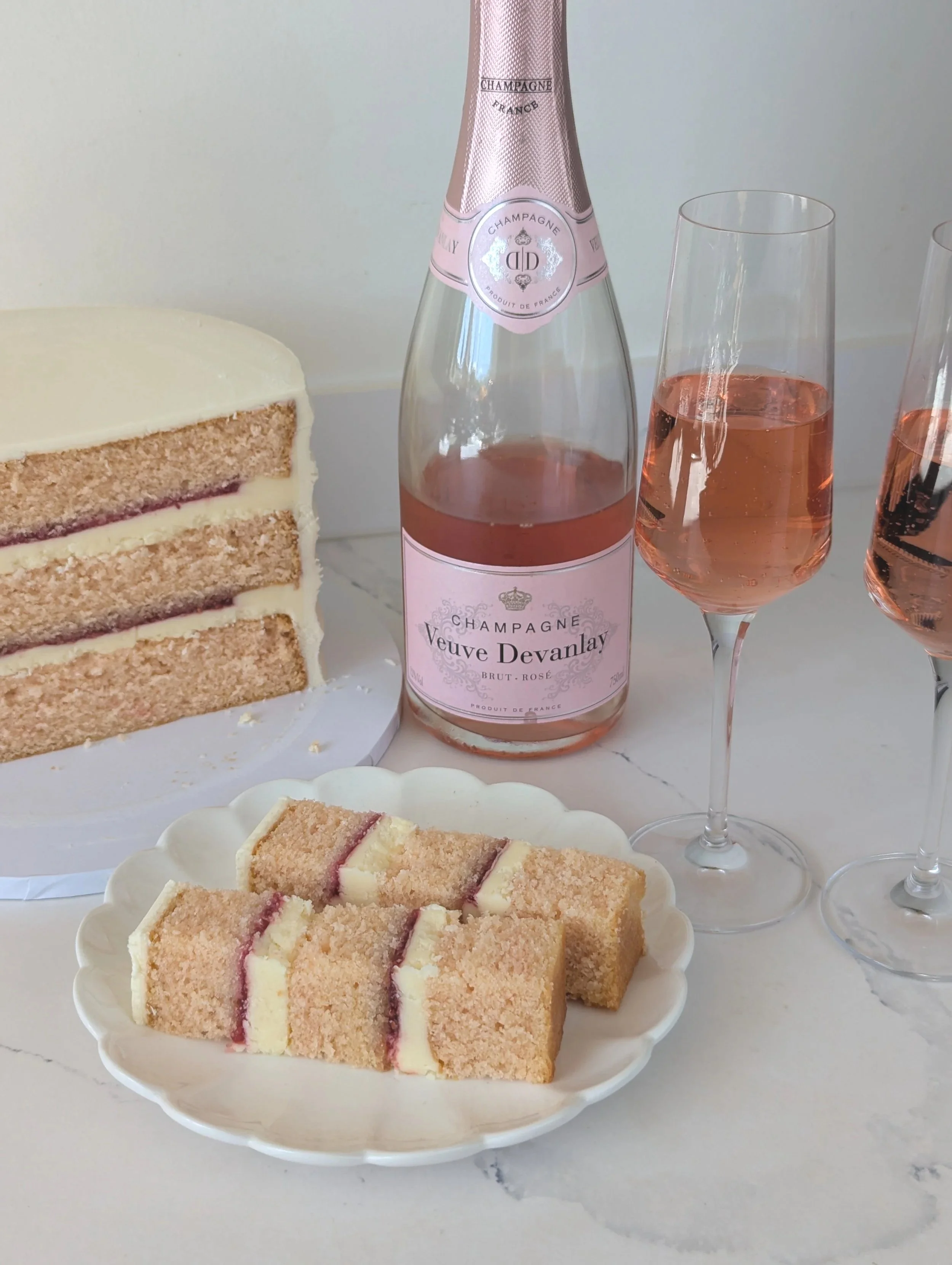 Slices of layered Pink Champagne cake with white icing and raspberry jam filling, a bottle of Veuve Devanlay rosé champagne, and two filled champagne flutes on a light-colored surface.