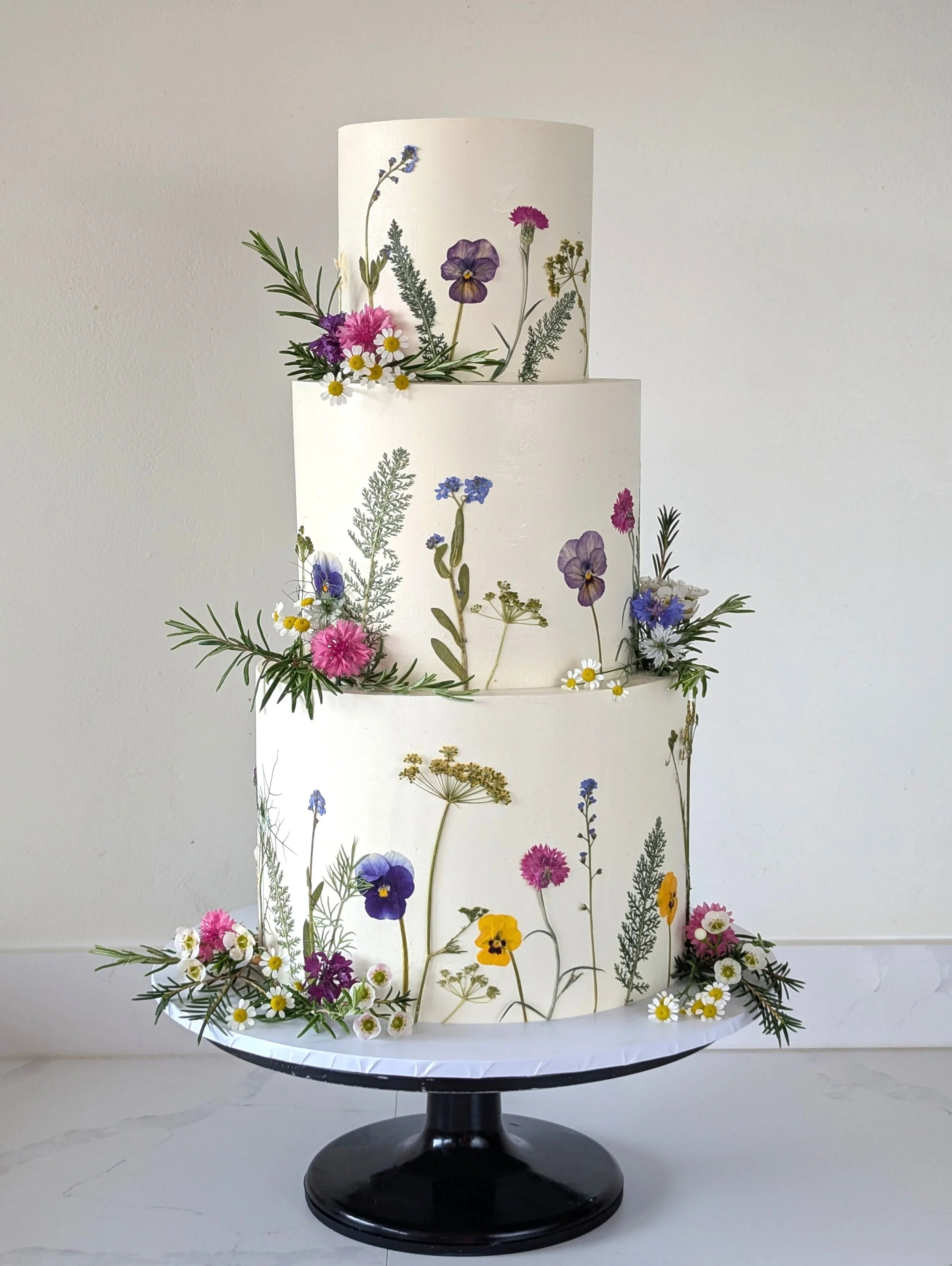 A tiered cake decorated with pressed and dried flowers, greenery, and daisies on a black cake stand with a plain white background.