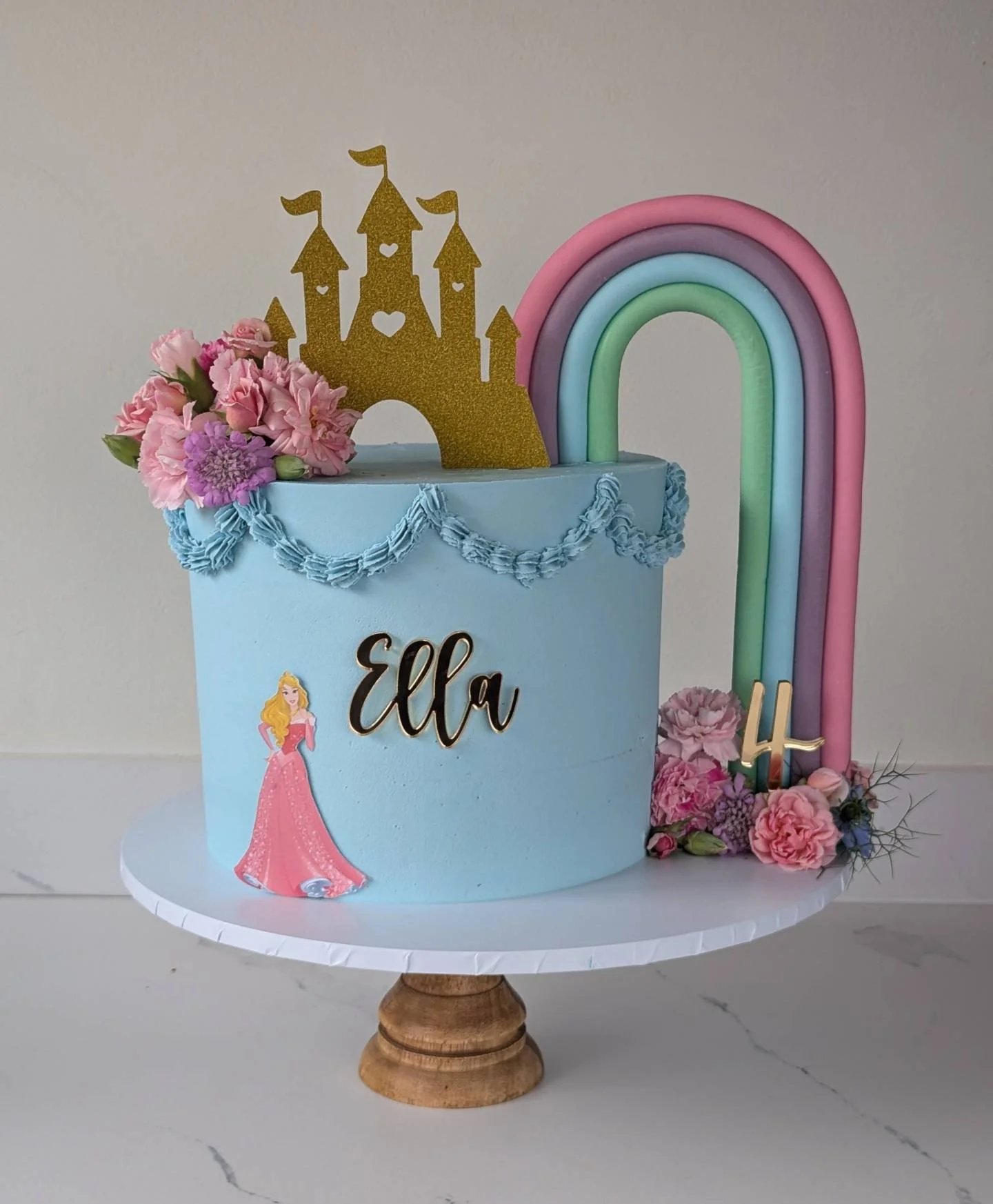 A children's birthday cake with a princess theme. The cake is light blue with pink and purple flowers, a gold castle topper, a rainbow decoration, and the name Ella written on the front.