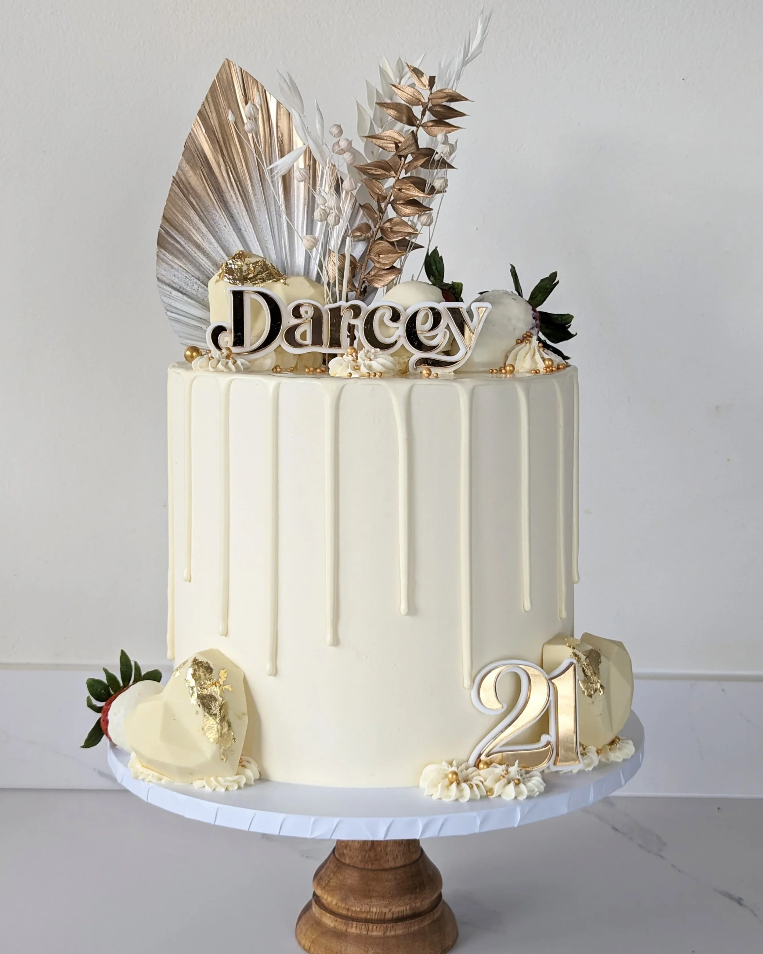 Elegant white cake decorated with gold accents, strawberries with gold leaf, and a gold "Darcy" cake topper, featuring gold and white dried foliage and a "2021" number sign, on a wooden cake stand.