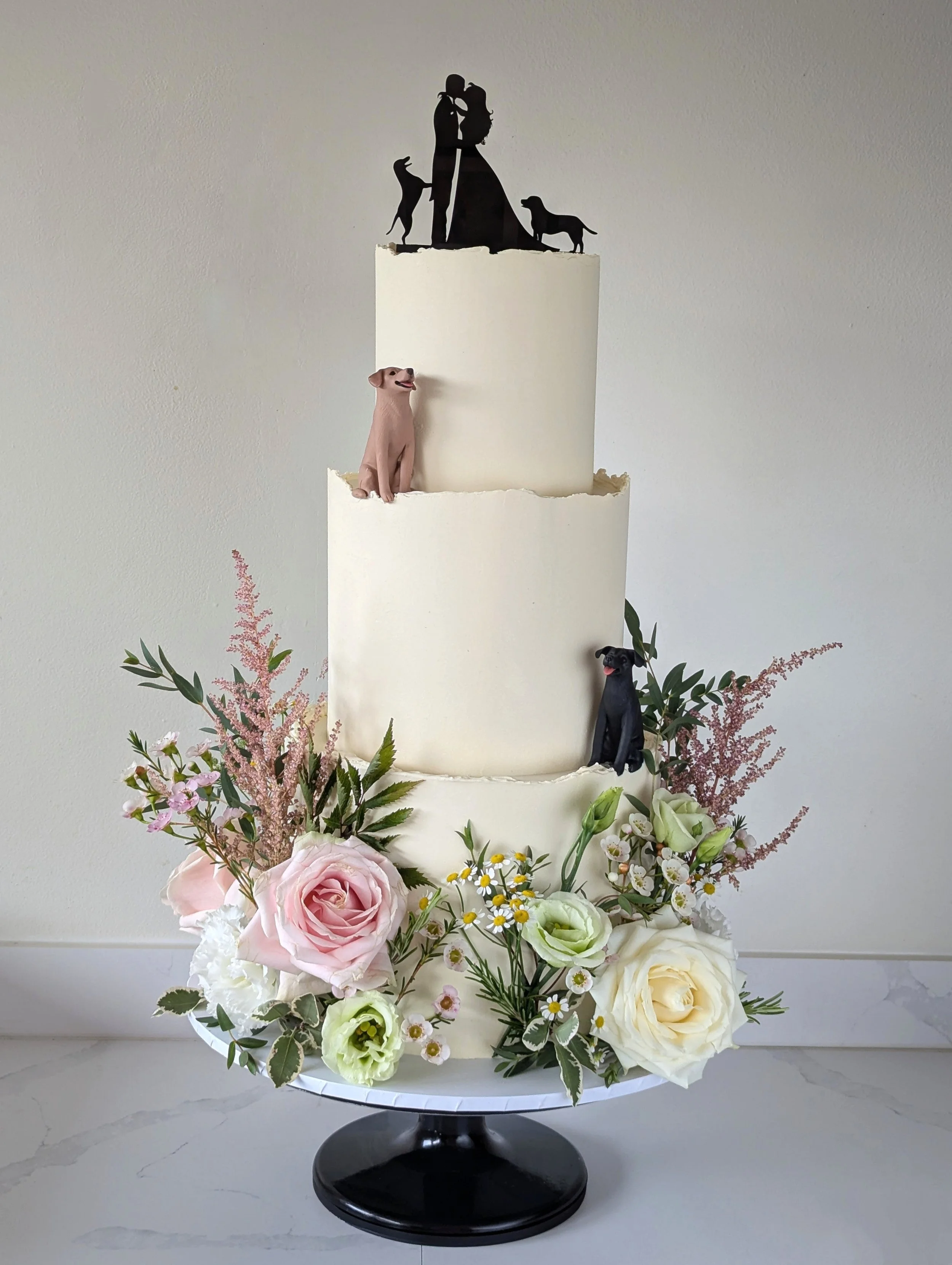 A floral cake with a black silhouette cake topper of a couple kissing and two dogs, decorated with pink, white, and green flowers, on a black cake stand. Syrencot, Wiltshire. 