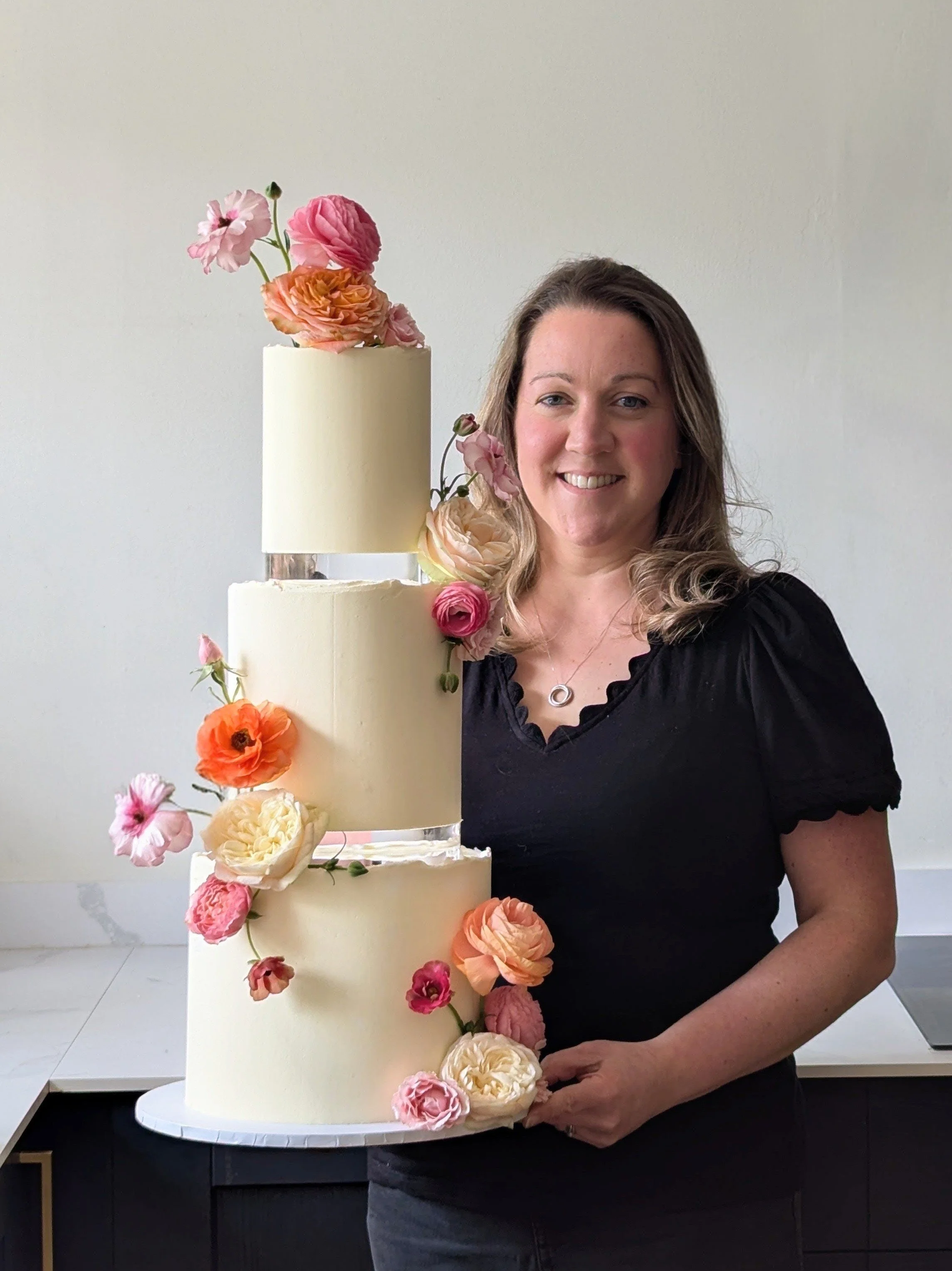 Jane (aka Mrs Wiltshire) with a three-tiered wedding cake decorated with pink, peach, and white flowers.