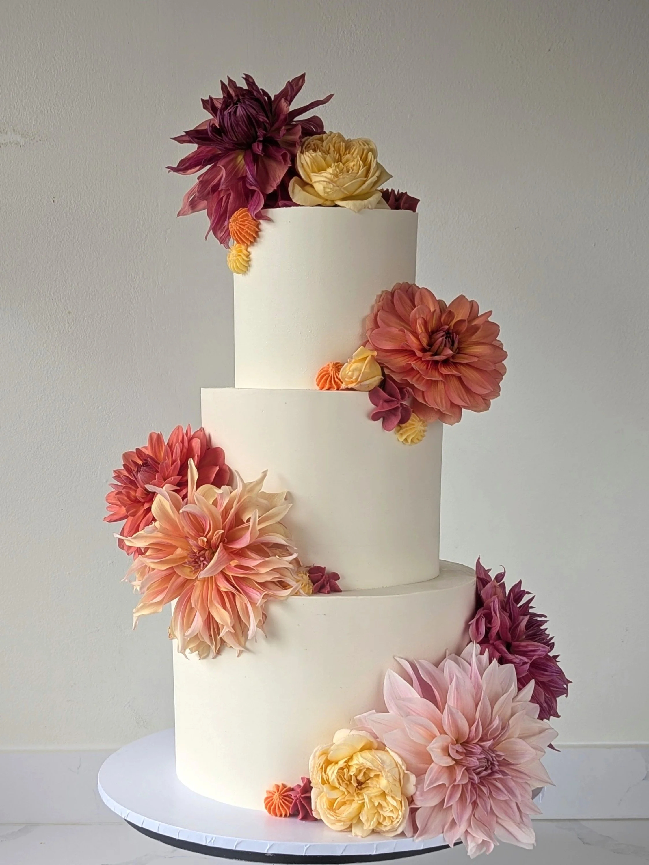 A white tiered cake decorated with large pastel-colored dahlias and smaller orange and pink flowers, with flowers cascading down the side.