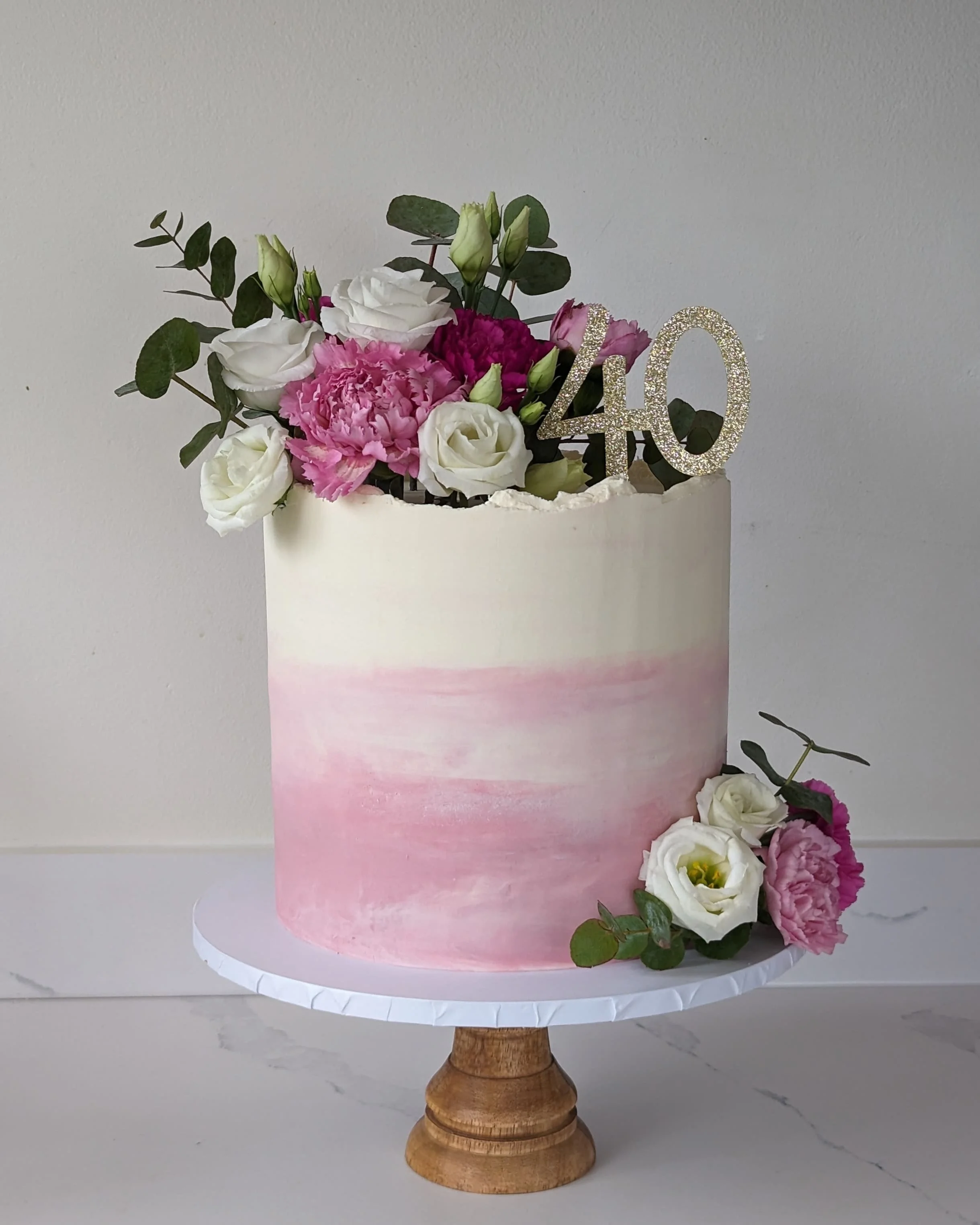 A pink ombre tiered cake on a wooden cake stand, decorated with a floral arrangement of white and pink flowers and green leaves, with a glittery '40' topper.