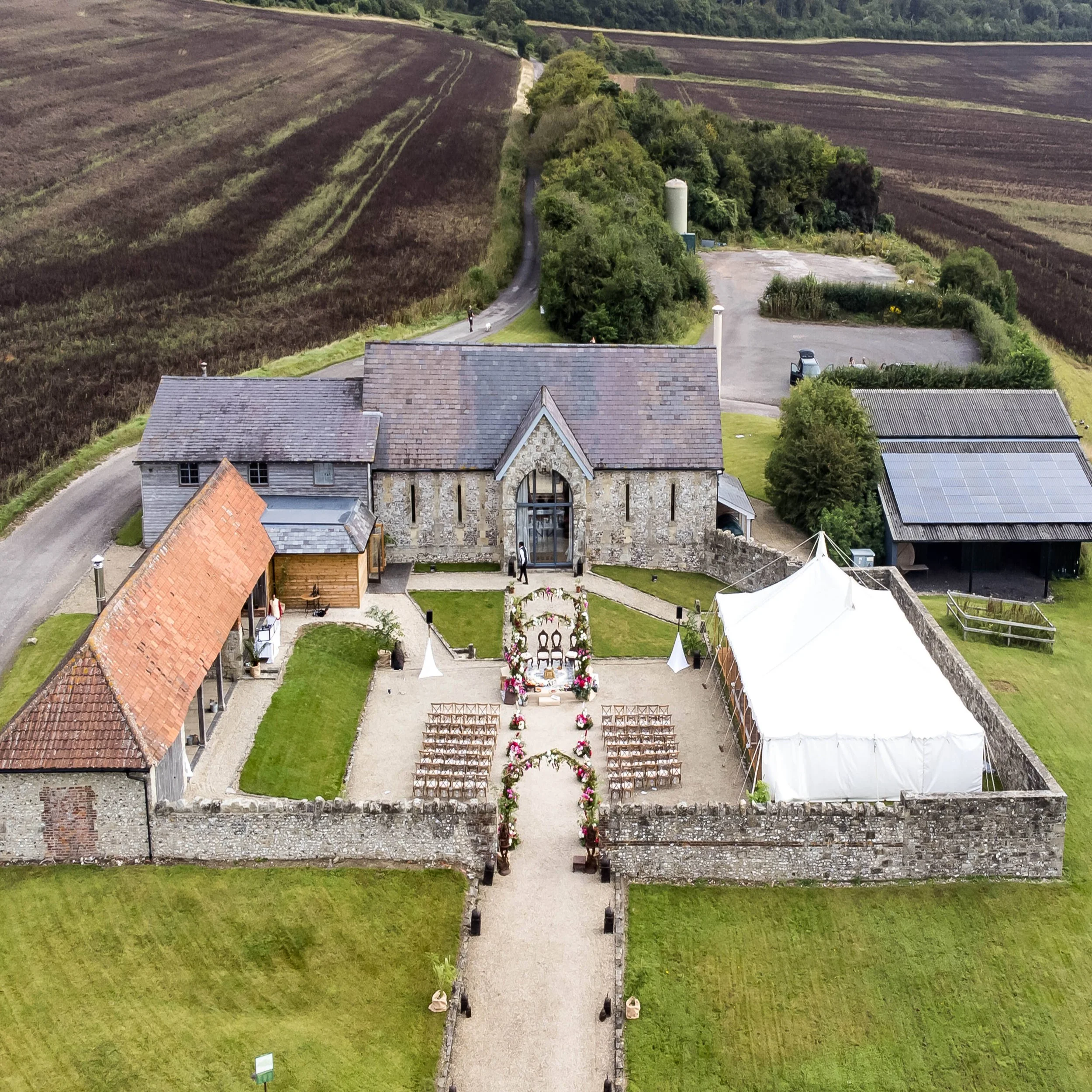Bake Barn, on the Fonthill Estate. A rural wedding setup on a gravel pathway in front of a stone and brick building, decorated with flowers and white tents, surrounded by green lawn and nearby parking area, with rolling farmland in the background.