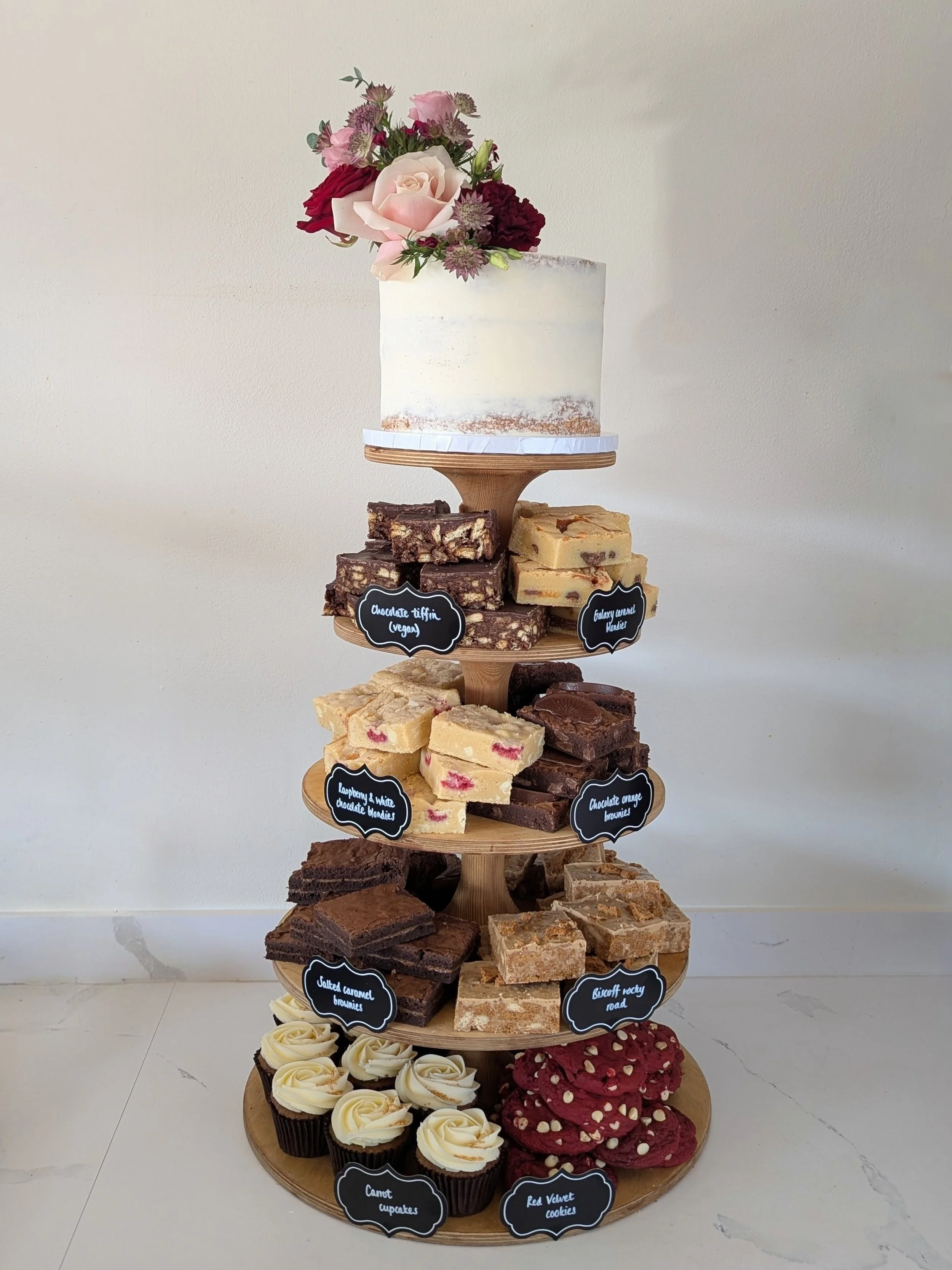 A multi-tiered dessert stand with various cakes and cookies, topped with a white frosted cake decorated with pink and red flowers. The tiers include chocolate tiffin, galaxy vortex blondies, raspberry & white chocolate blondies, chocolate orange brow