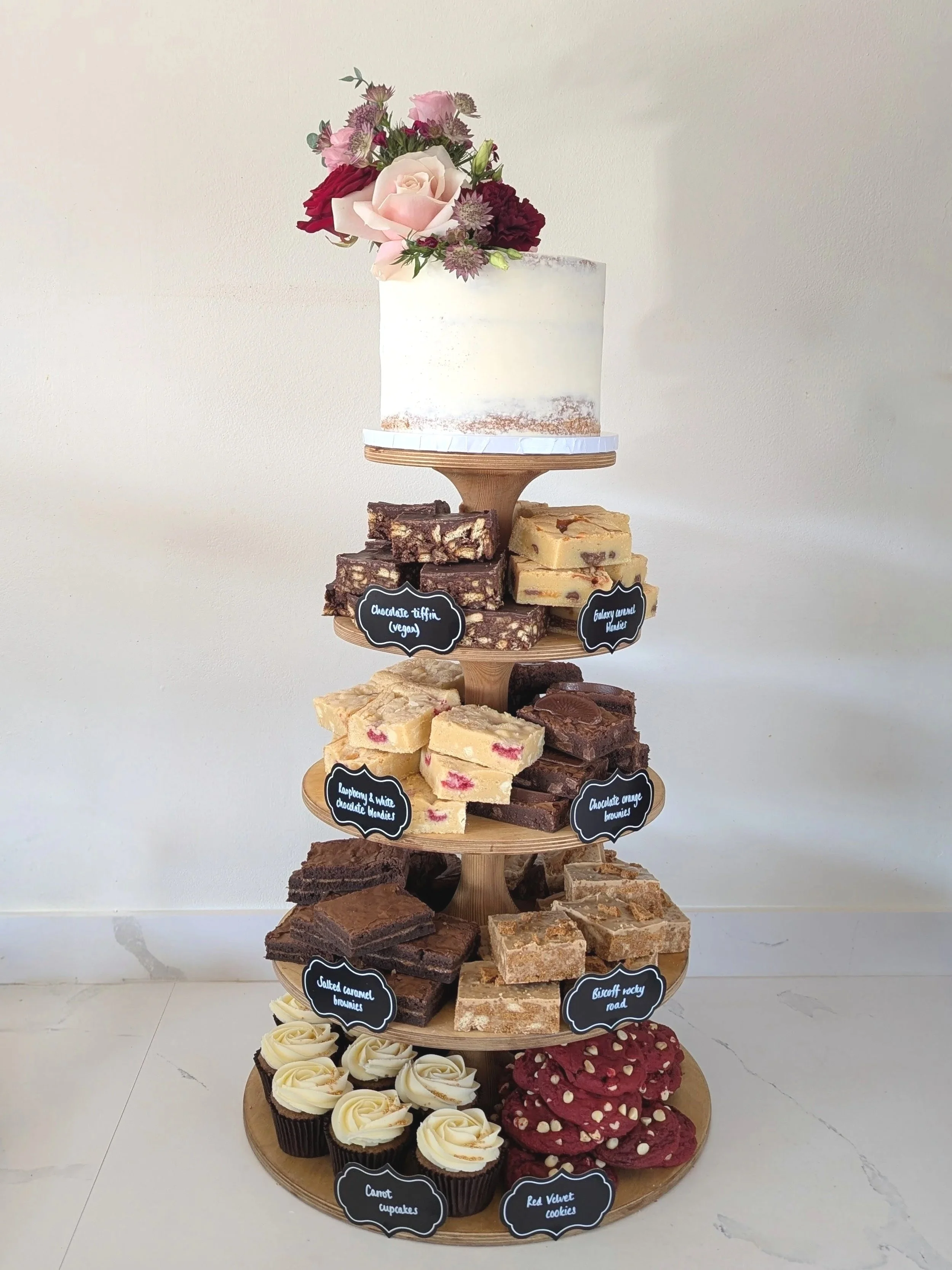 A multi-tiered display stand with various desserts, including cupcakes, brownies, and bars, topped with a white cake decorated with purple and pink flowers, and labeled with small chalkboard signs. Delivered to Elmhay Park.