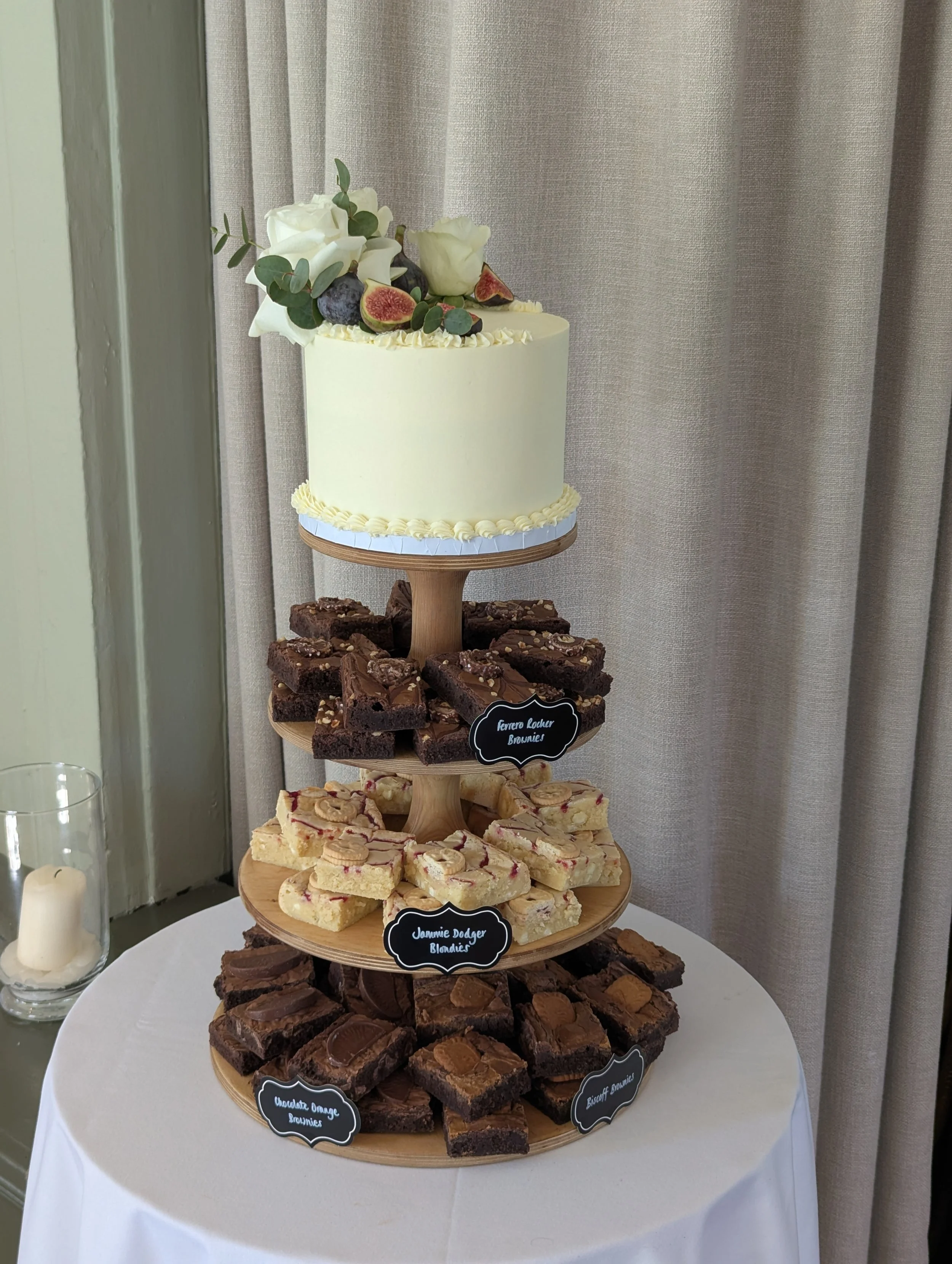 Tiered dessert display with assorted brownies and blondies, each type labeled with small black chalkboard signs, topped with a small cake decorated with white flowers, figs, blueberries, and greenery. Wedding cake in Bath. 