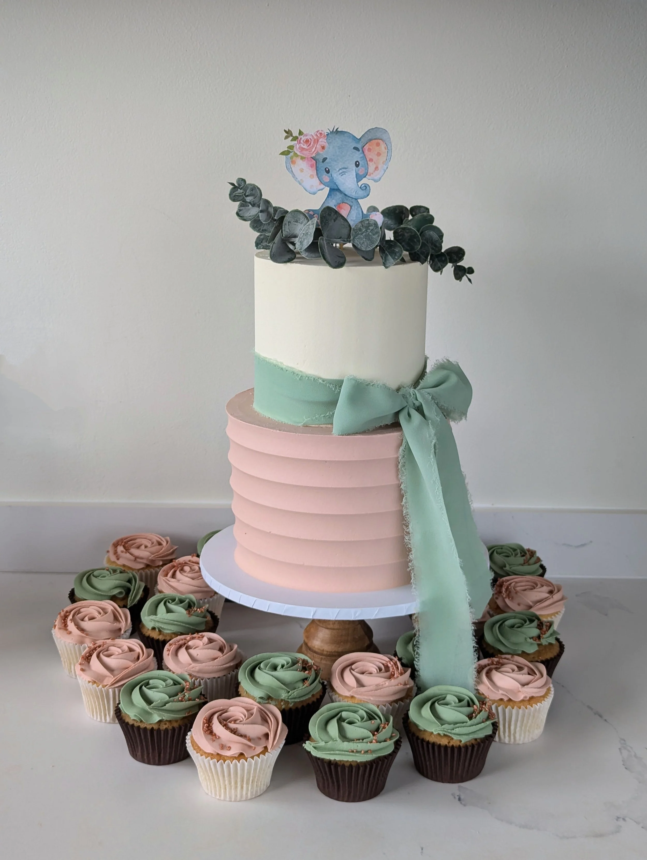 A tiered cake decorated with a paper elephant figurine with pink flowers on its head on top, surrounded by black and pastel pink cupcakes with swirl frosting, on a white stand and surrounded by flower-shaped cupcakes with green, pink, and brown frost