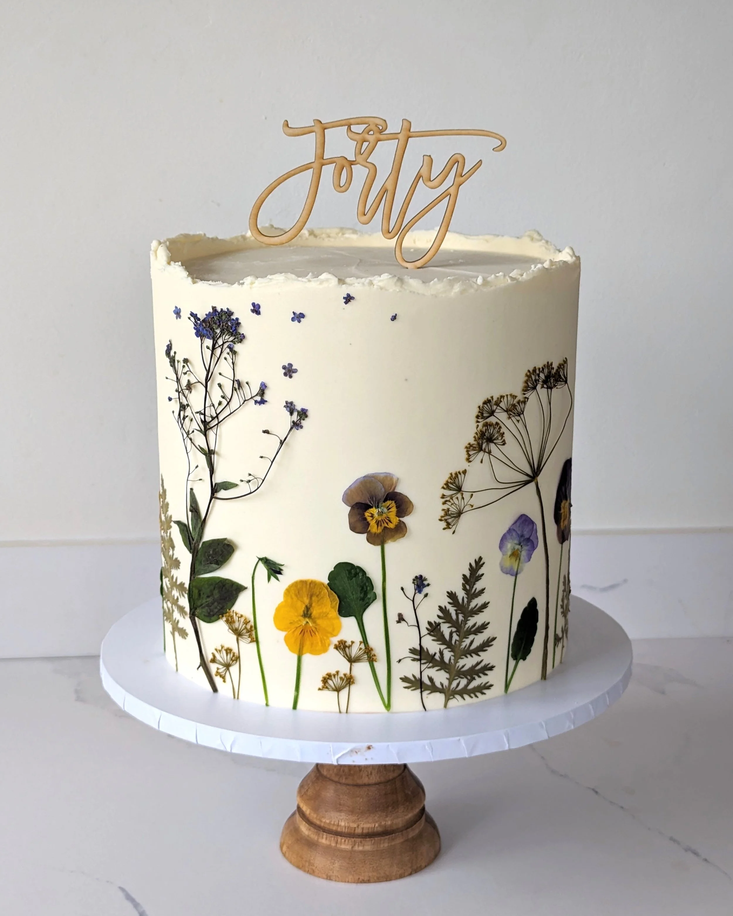 A decorated cake with a botanical theme on a white cake stand. It features dried flowers and pressed plants with a 'Joy' topper.