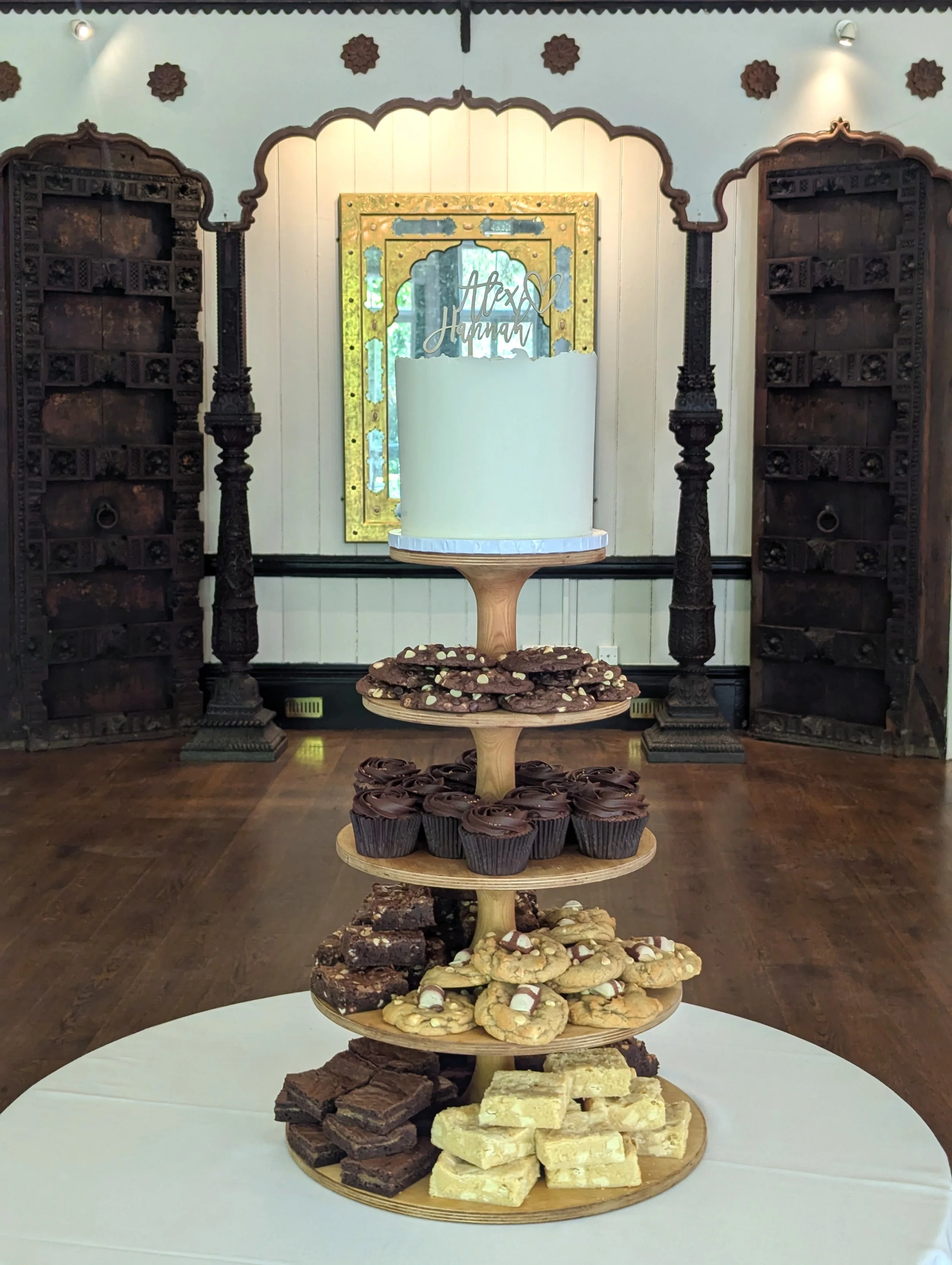 Tiered display of assorted cookies and brownies on wooden plates with a decorative cake stand in the center, in a room with ornate woodwork and a mirror in the background. Larmer Tree wedding cake. 