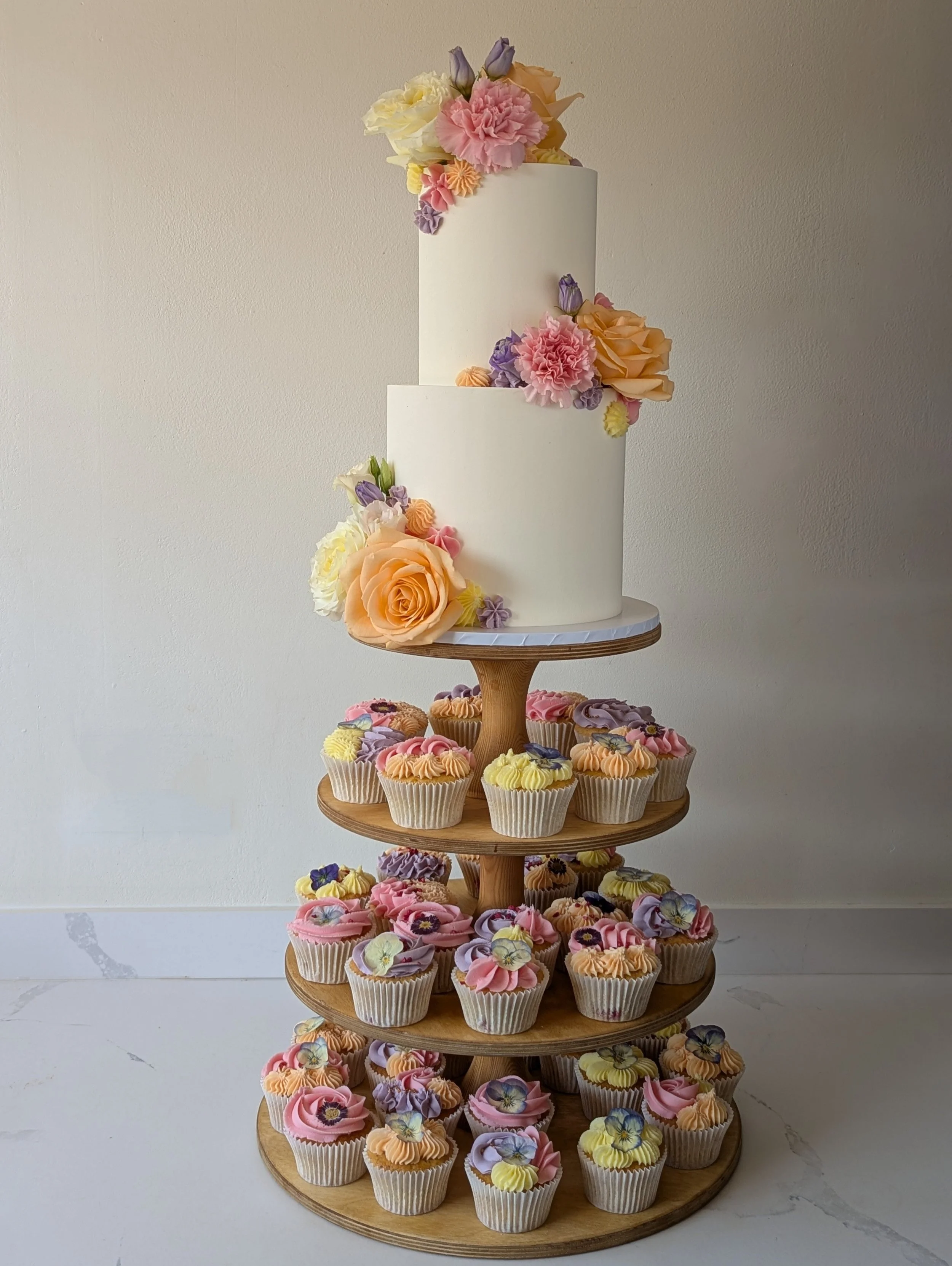 A tiered wooden cupcake stand with four levels, decorated with colorful paper cupcakes and topped with a white cake adorned with pastel-colored flowers.