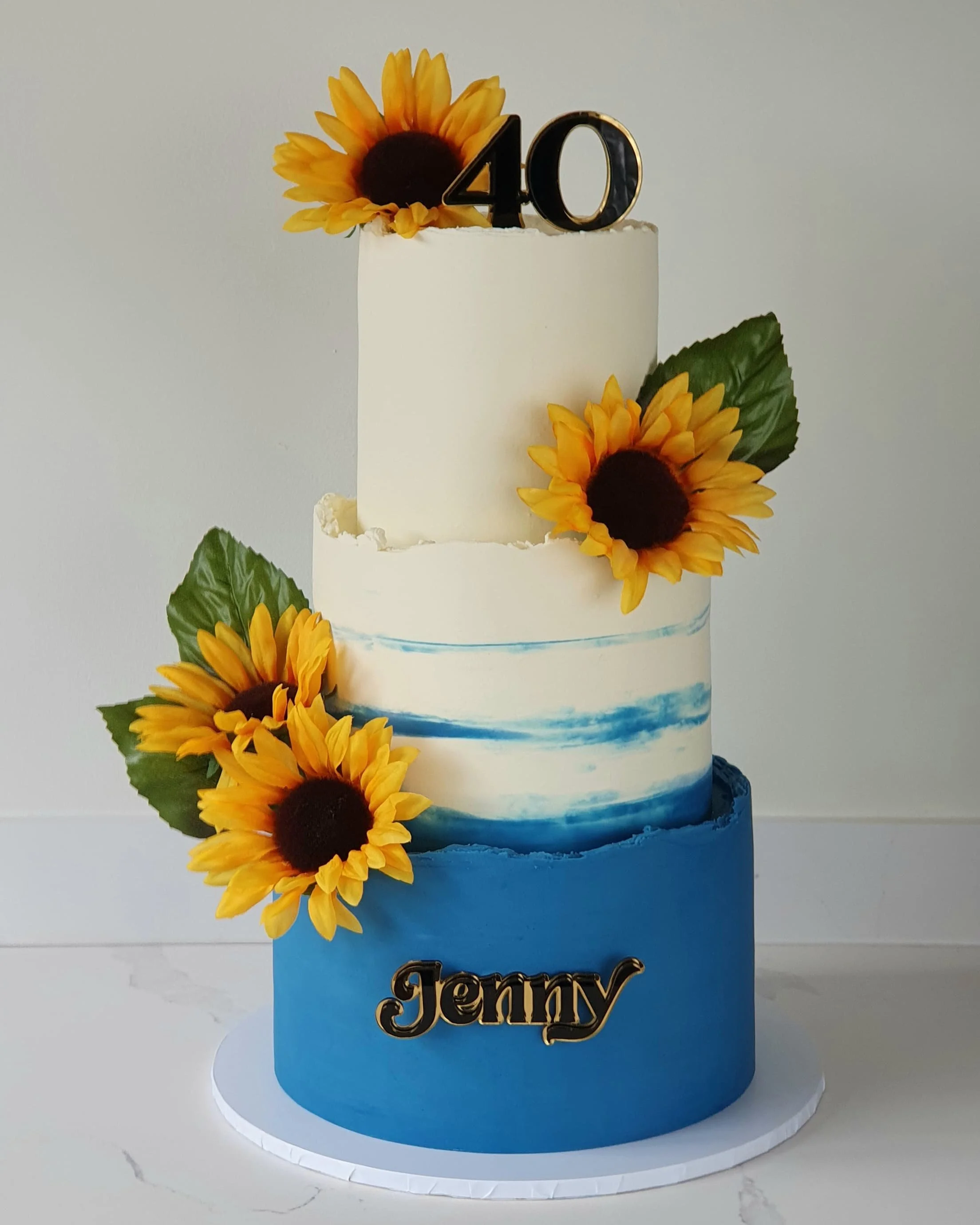 A three-tiered cake decorated with sunflowers and green leaves, featuring a black and gold '40' topper and the name 'Jenny' in gold at the bottom, against a plain white background.