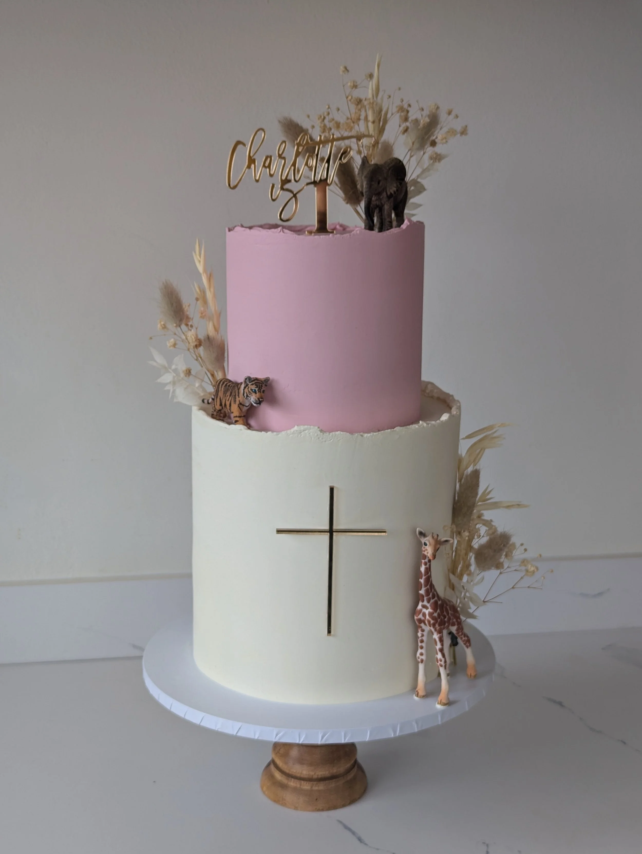 A white tiered cake with a pink top tier decorated with a gold 'Charlotte' topper, surrounded by small plastic animal figurines of a tiger, zebra, and an elephant, and dried beige and white floral arrangements.