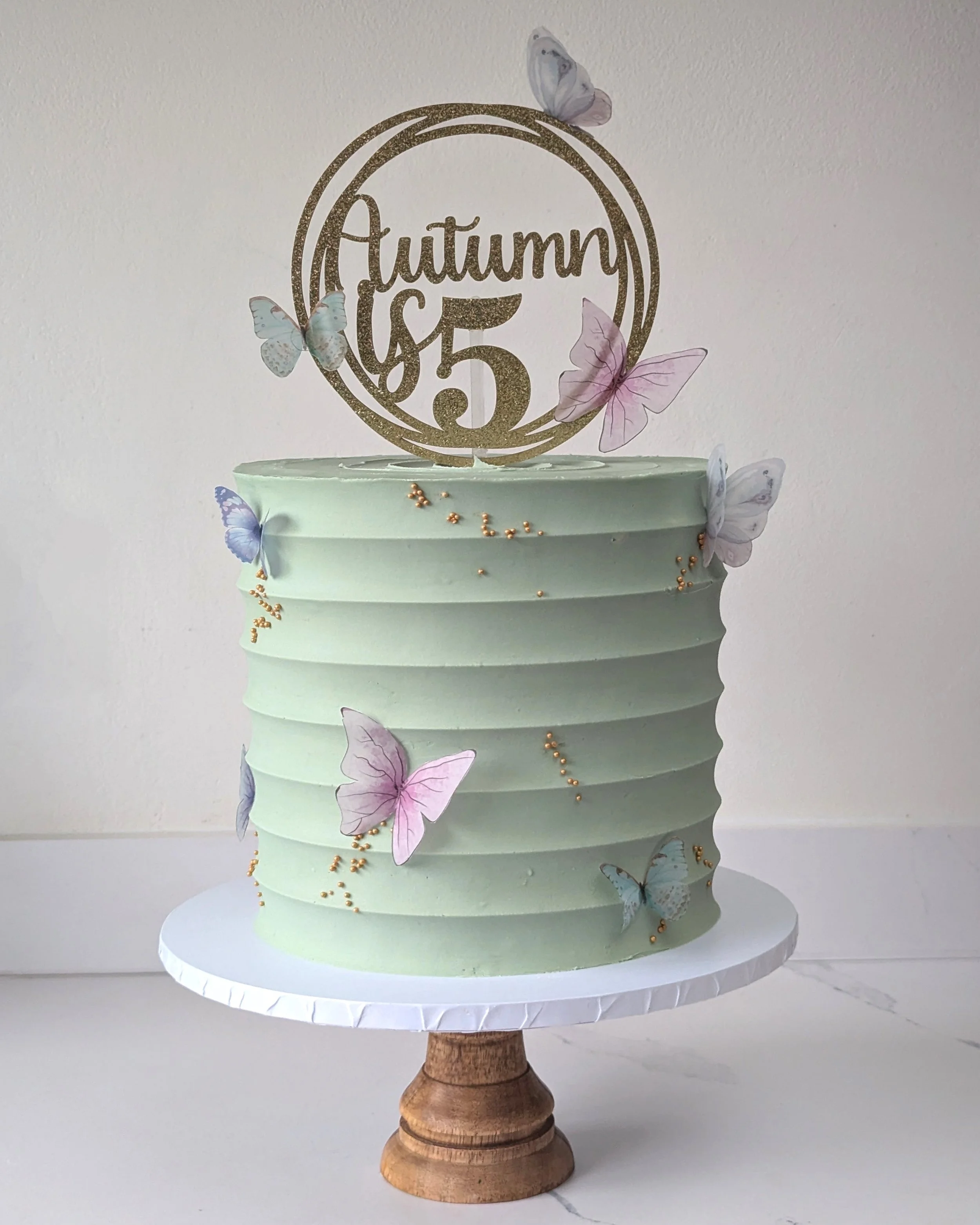 A cake with a multi-tiered design, decorated with butterfly embellishments and small golden beads, featuring a circular topper that reads 'Autumn 5,' and displayed on a wooden cake stand.