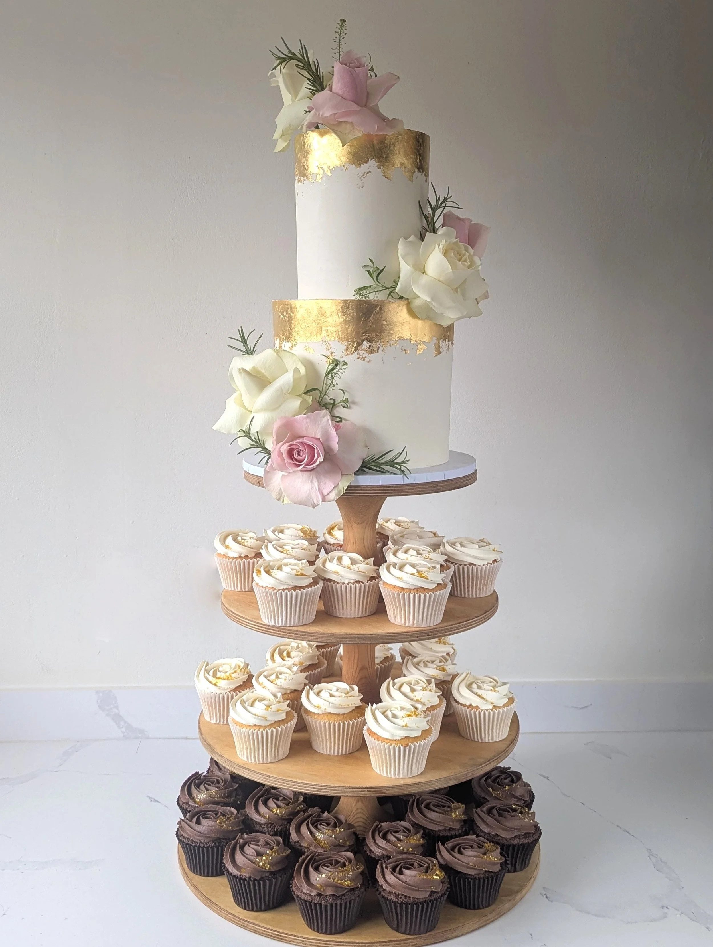 A multi-tiered dessert stand with cupcakes, decorated for a celebration. The top tier features a white and gold cake adorned with pink and white roses and greenery. The middle tier has cupcakes with white frosting and gold accents, and the bottom tie