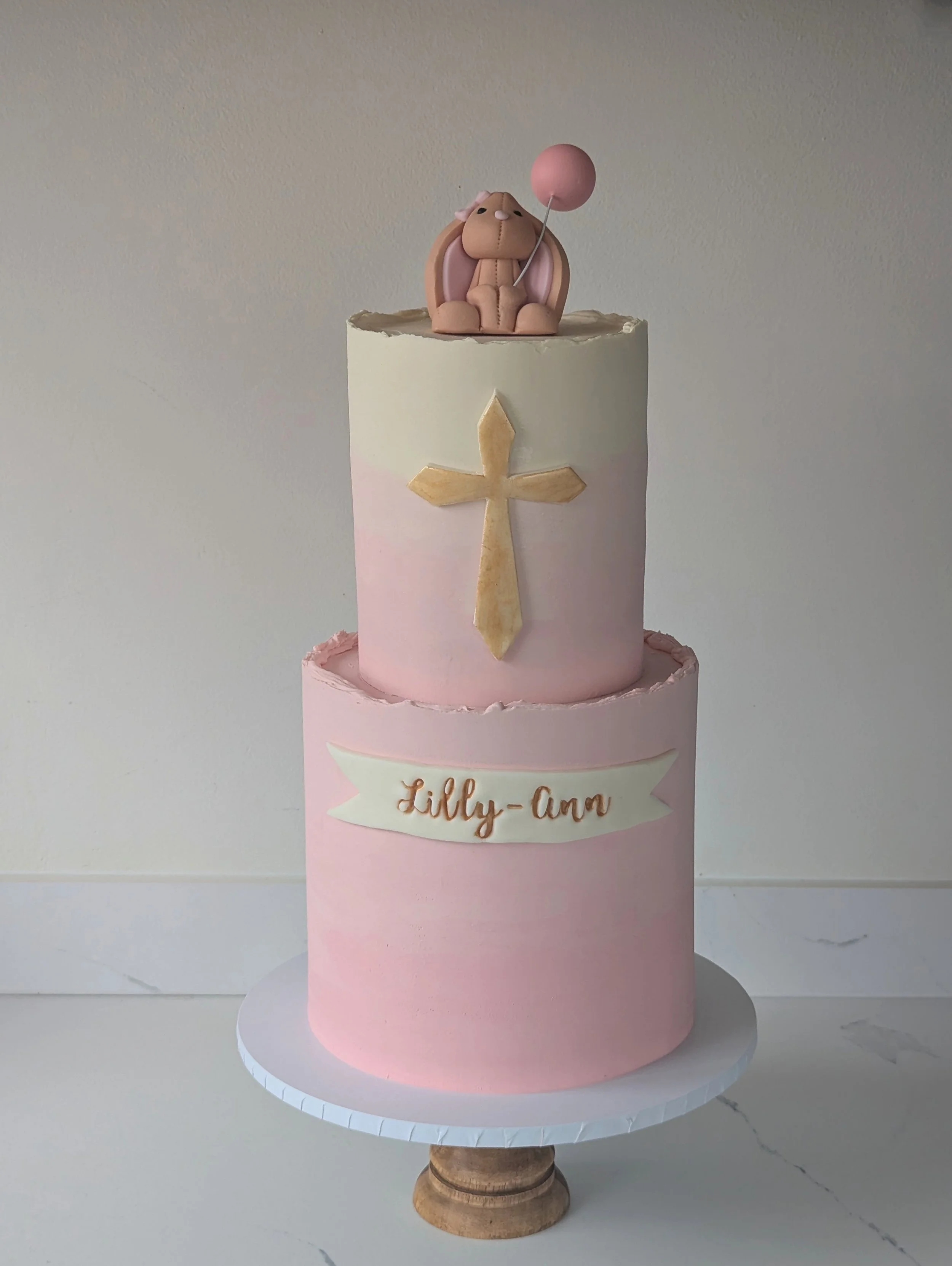 A multi-tiered cake with pink and white icing, a cross decoration on the front, and a fondant baby lion figure holding a pink balloon on top.