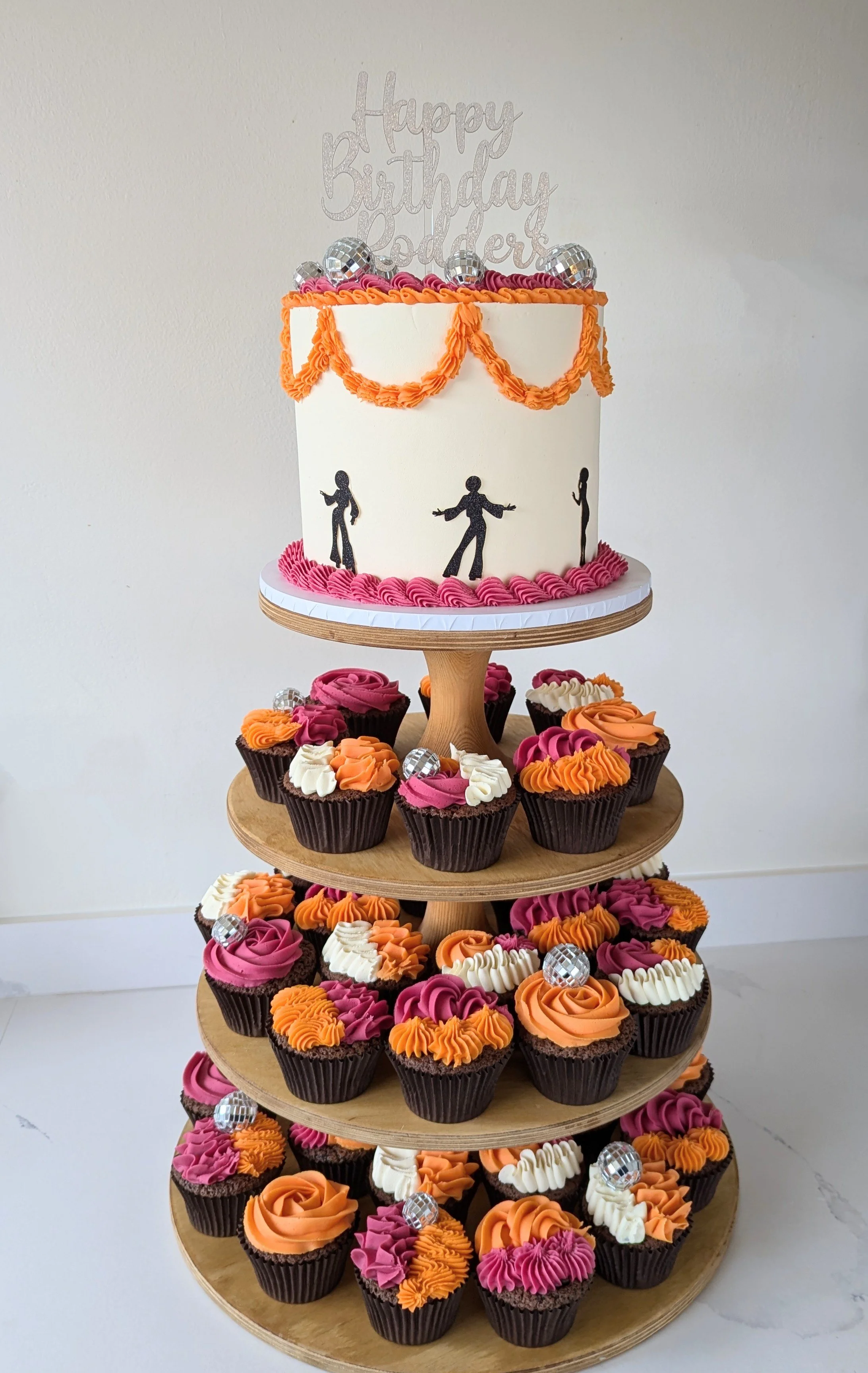 A tiered birthday cake stand decorated with cupcakes and a white cake on top. The cake has orange piping and black silhouettes of dancers, pink frosting at the base, and a topper that says 'Happy Birthday' with silver disco balls and purple decoratio
