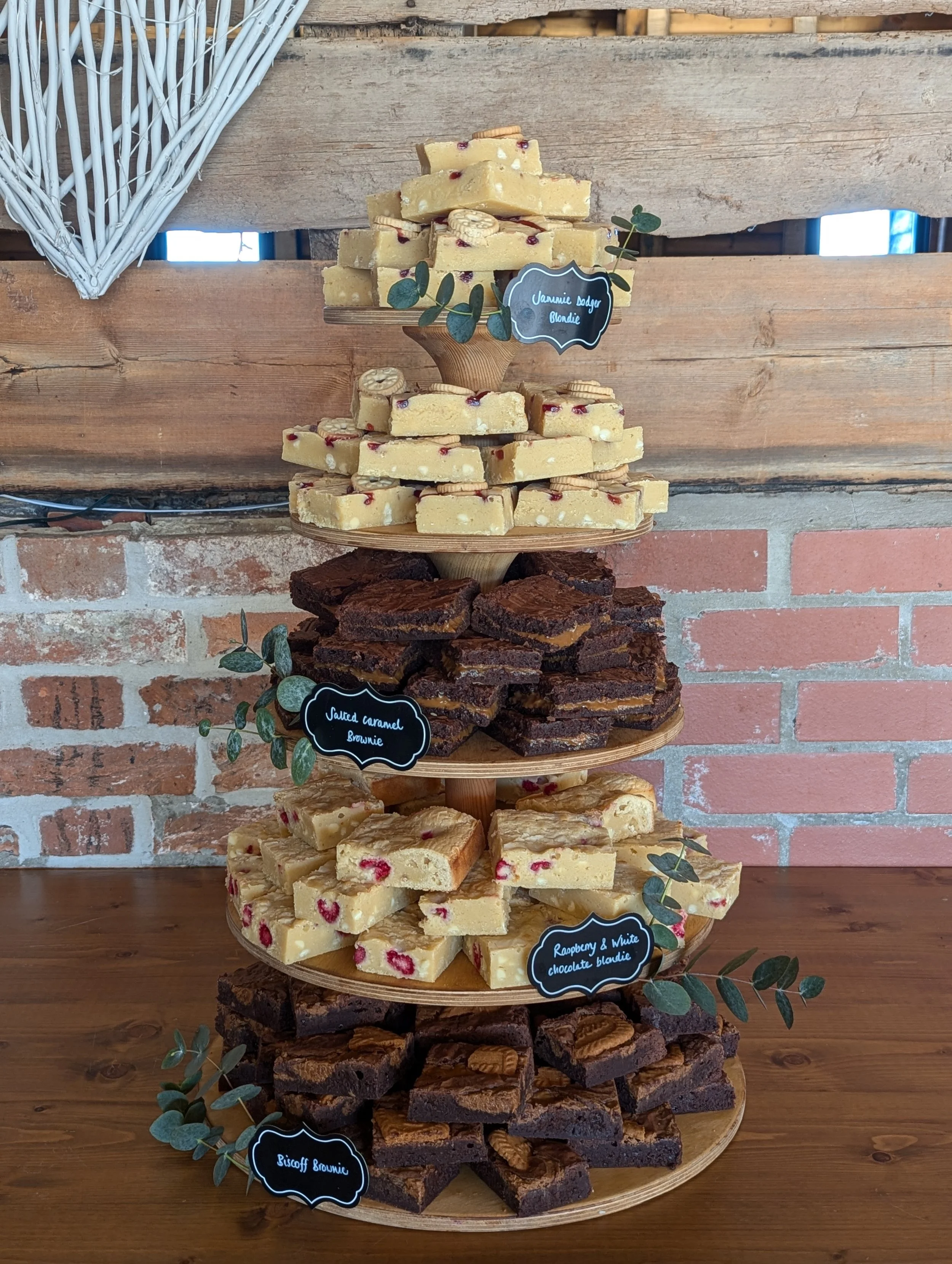 A multi-tiered display stand with various types of white and chocolate blondies, each labeled with handwritten chalkboard signs, set against a rustic brick and wood background.