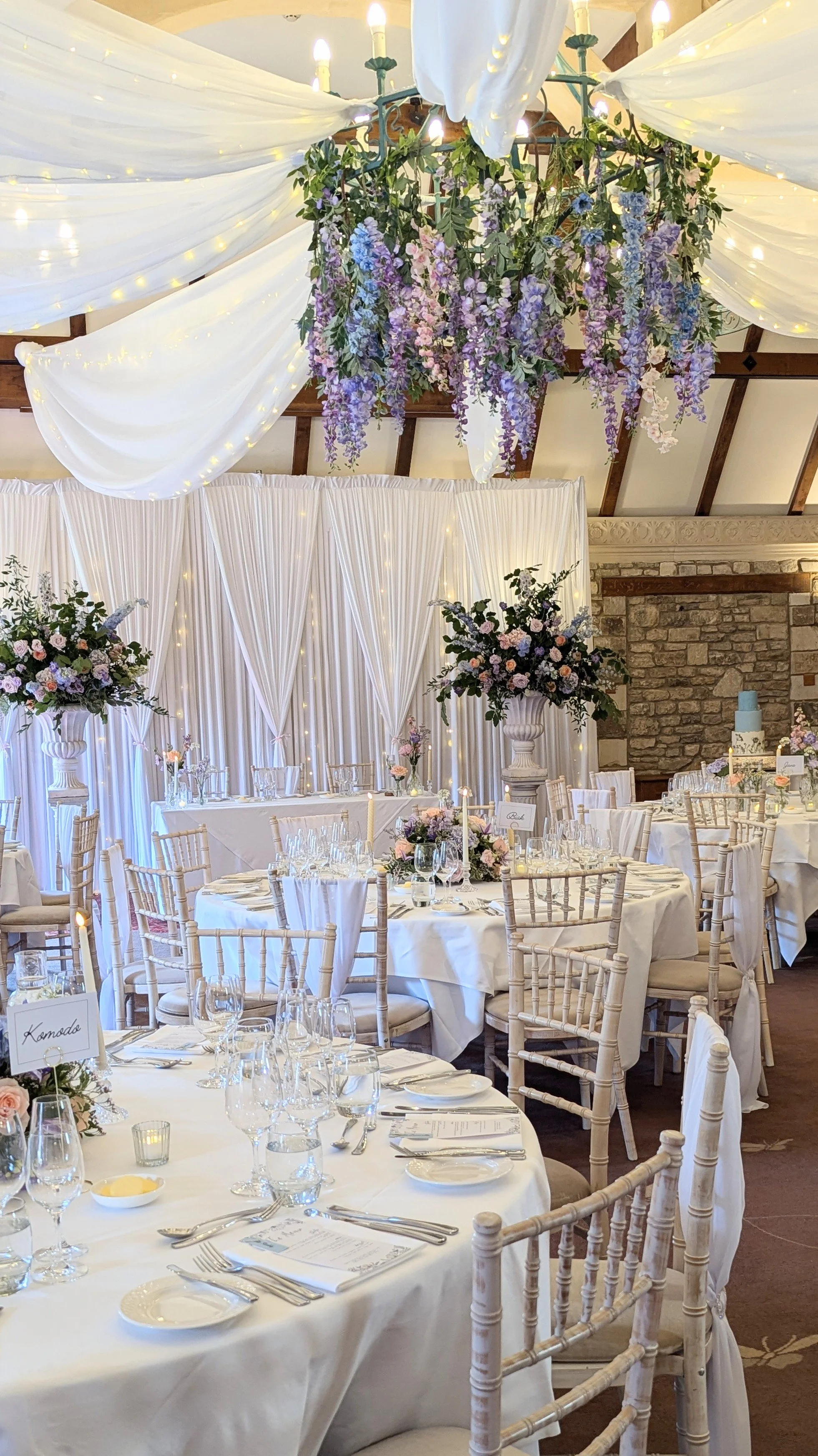 A beautiful wedding cake at the Manor House, Castle Combe. Elegant banquet hall decorated with white drapes, fairy lights, and floral arrangements in tall vases.
