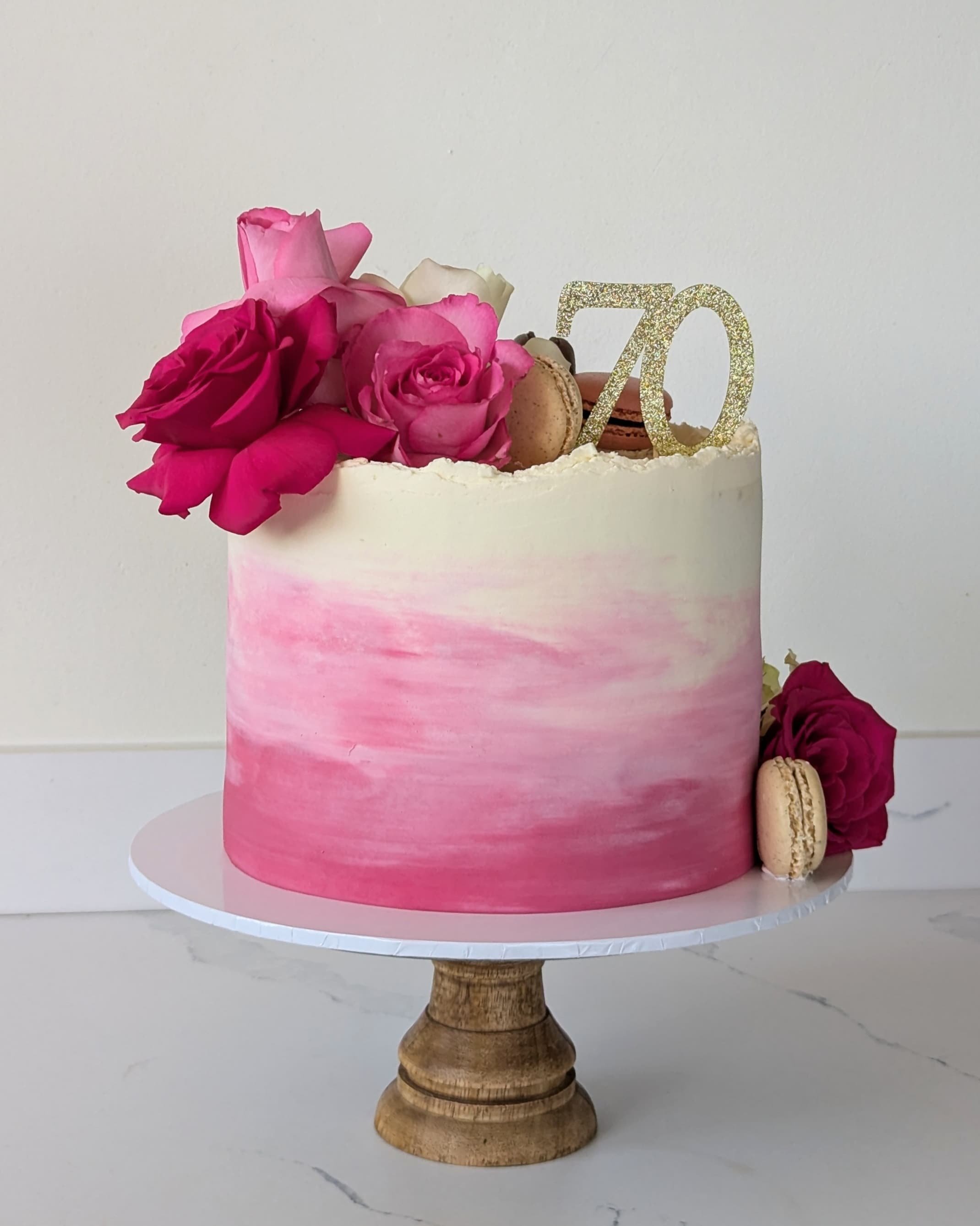 A pink and white ombre cake decorated with pink and white roses, macarons, and a gold glitter "70" cake topper, displayed on a wooden cake stand.