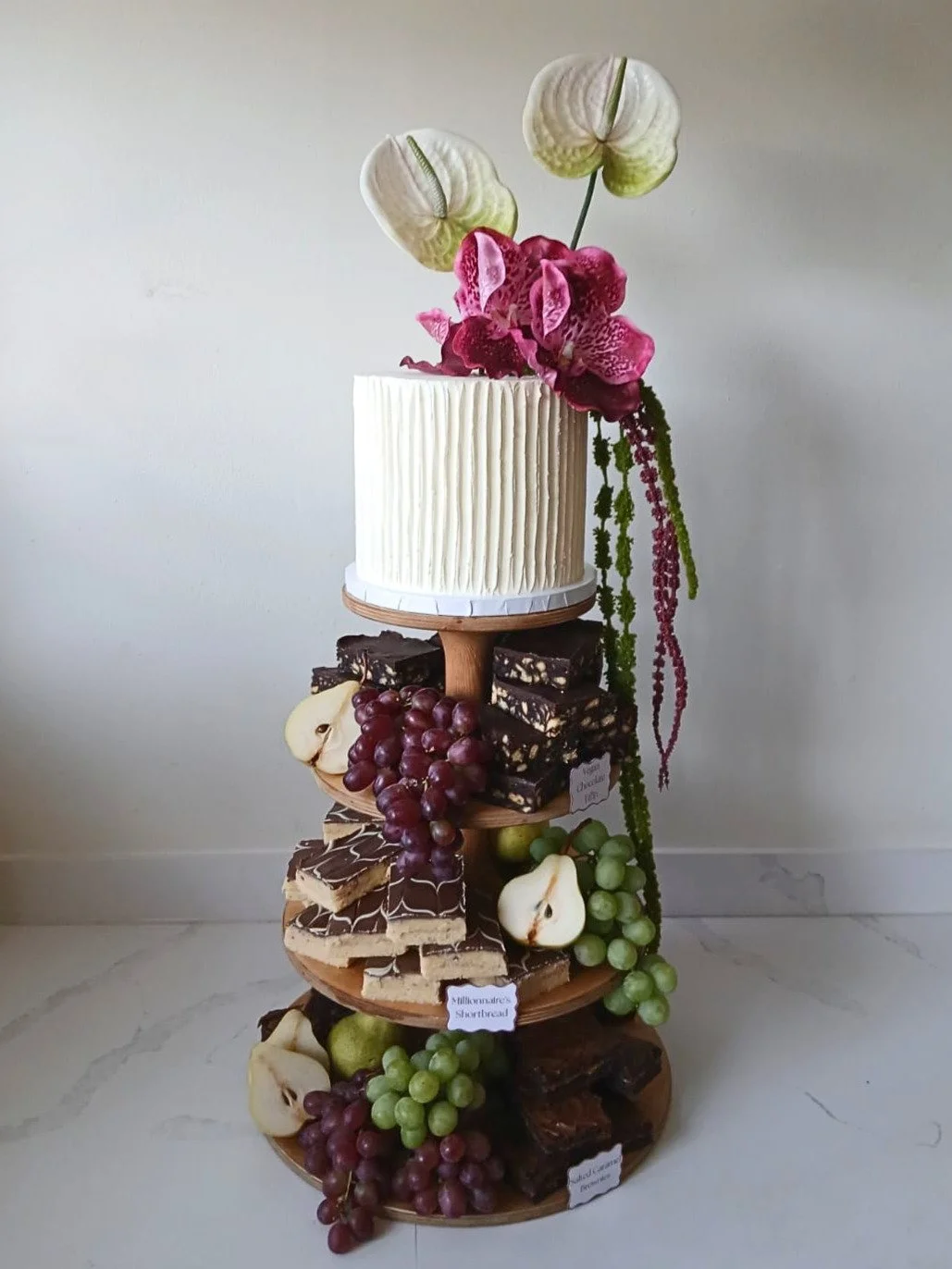 A multi-tiered dessert stand with a white frosted cake on top, decorated with an arrangement of pink and white orchids and anthuriums, and assorted chocolates, grapes, pears, and other fruits on the tiers below.