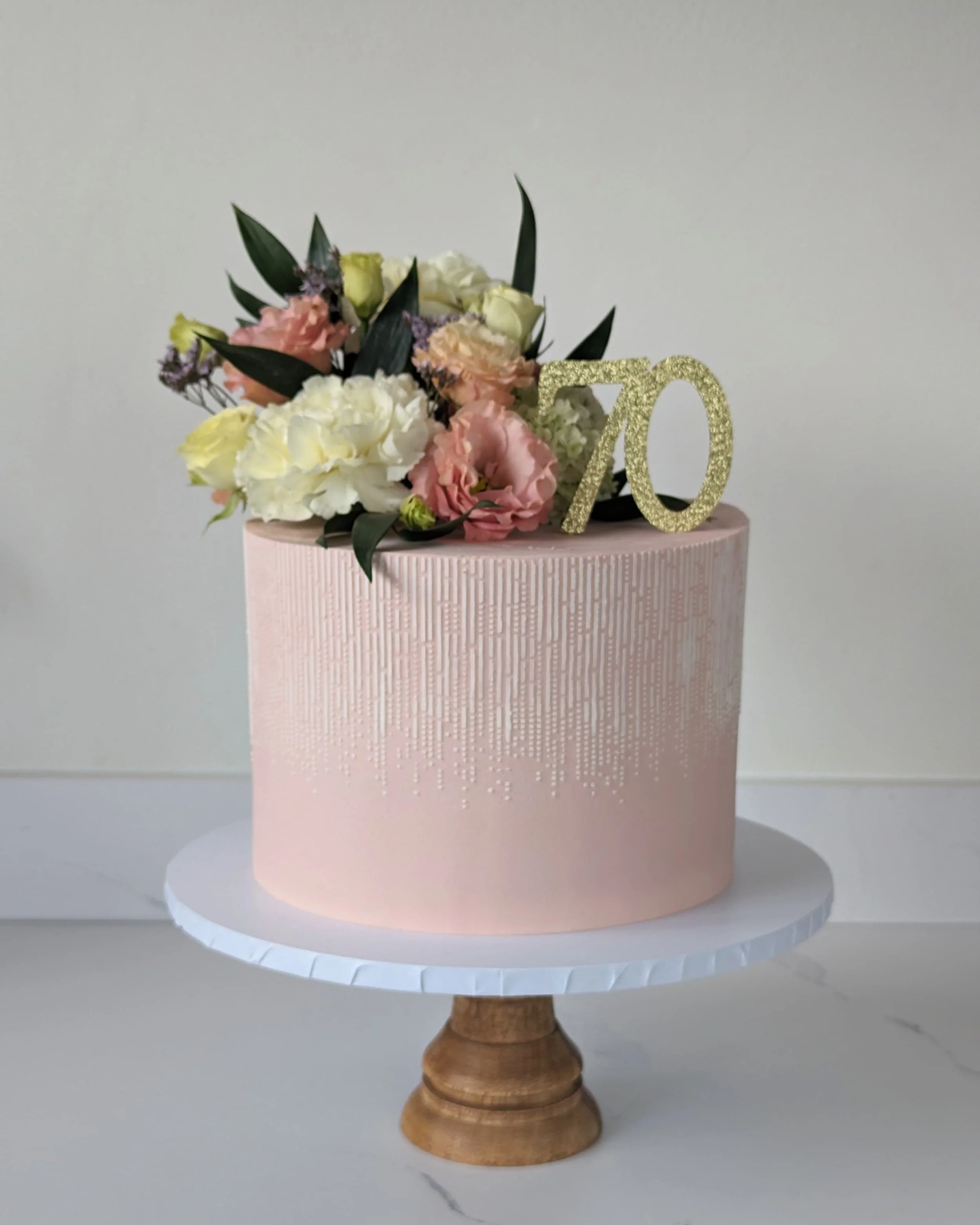 A round pink cake stand with a textured surface holds a bouquet of pink, white, and purple flowers, along with green leaves, and a gold glittery number '70' decoration. The cake stand is on a white surface against a plain white background.