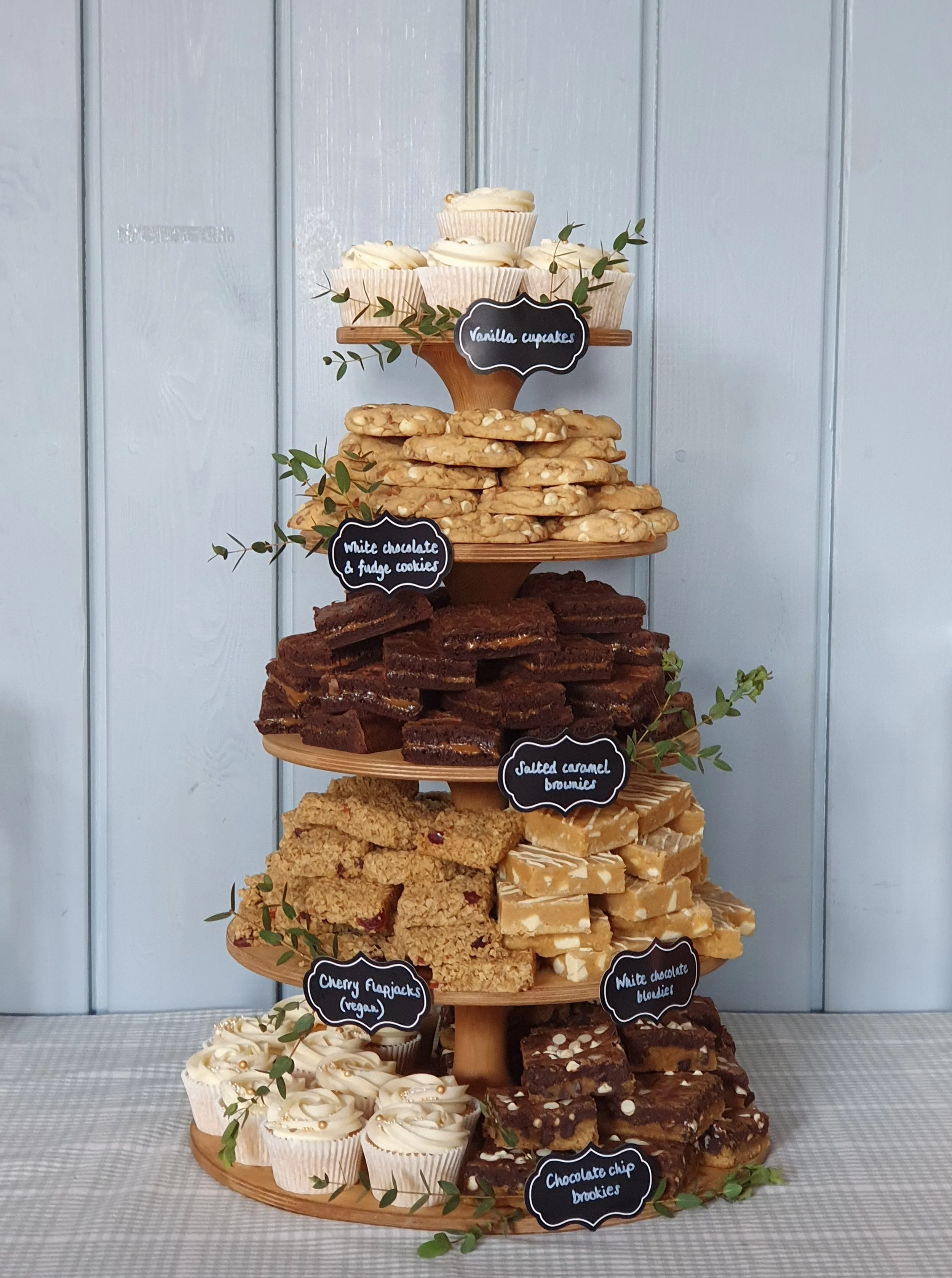 A multi-tiered dessert display with various baked goods and labels, including vanilla cupcakes, white chocolate & fudge cookies, salted caramel brownies, cherry flapjacks (vegan), white chocolate blondies, and chocolate chip cookies, decorated with g