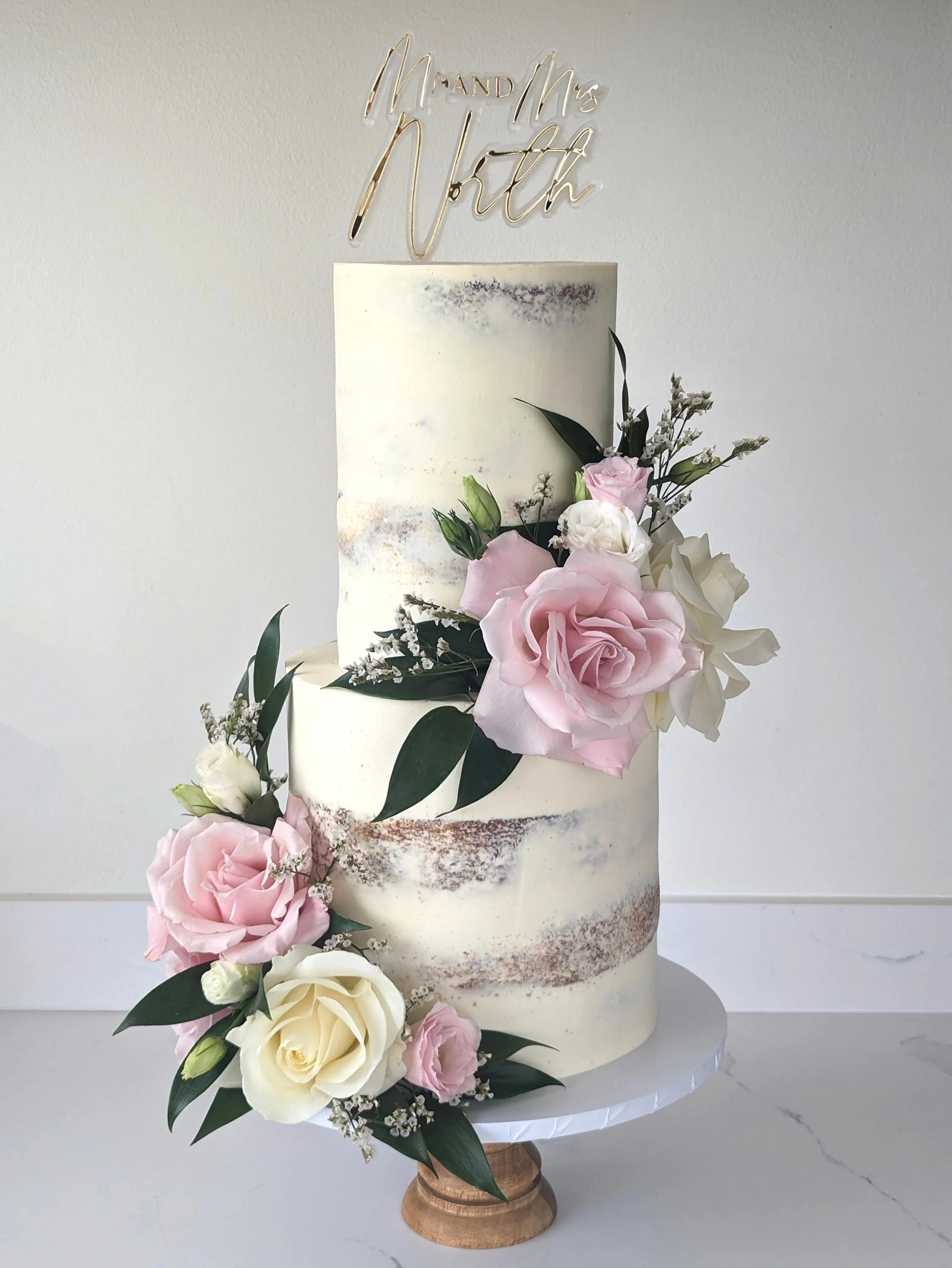 A multi-tiered wedding cake decorated with fresh pink and white roses, white filler flowers, and greenery, with a gold "Mr and Mrs North" cake topper on a marble stand.