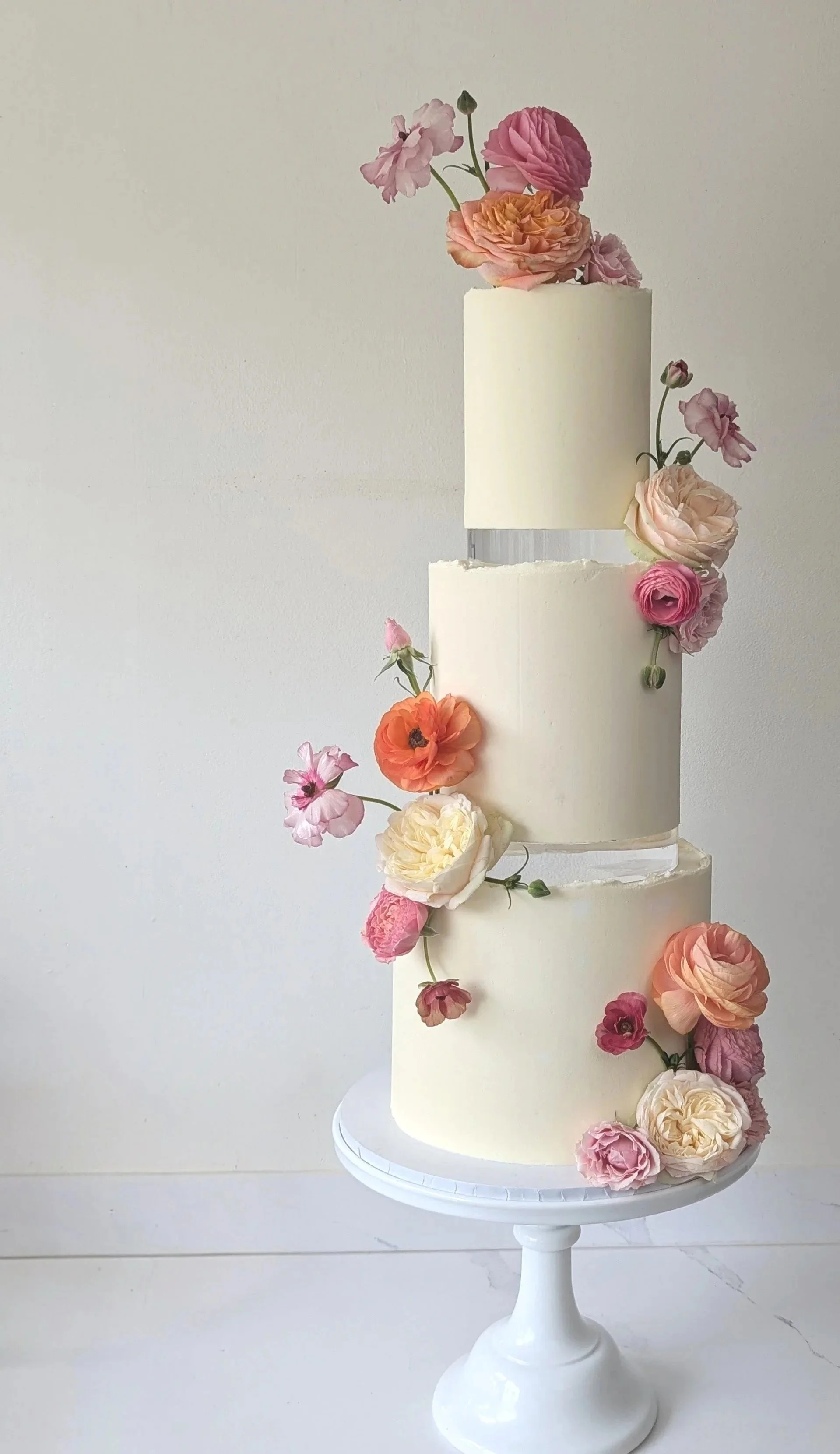 A three-tiered white wedding cake decorated with pink, peach, cream, and purple fresh flowers, placed on a white stand against a plain wall.