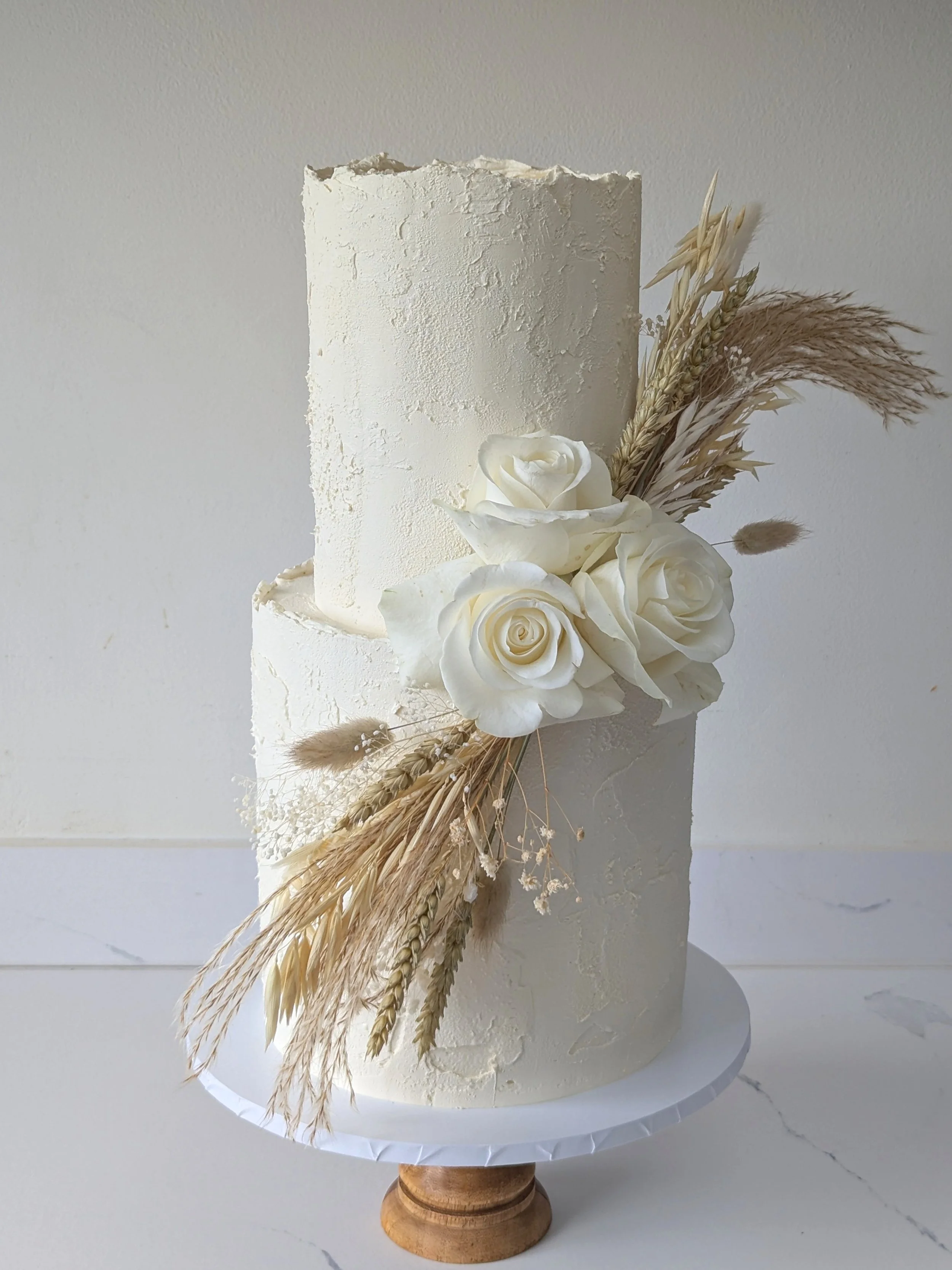 A white textured cake decorated with white roses, dried grasses, and pampas plumes, displayed on a white cake stand against a plain background.
