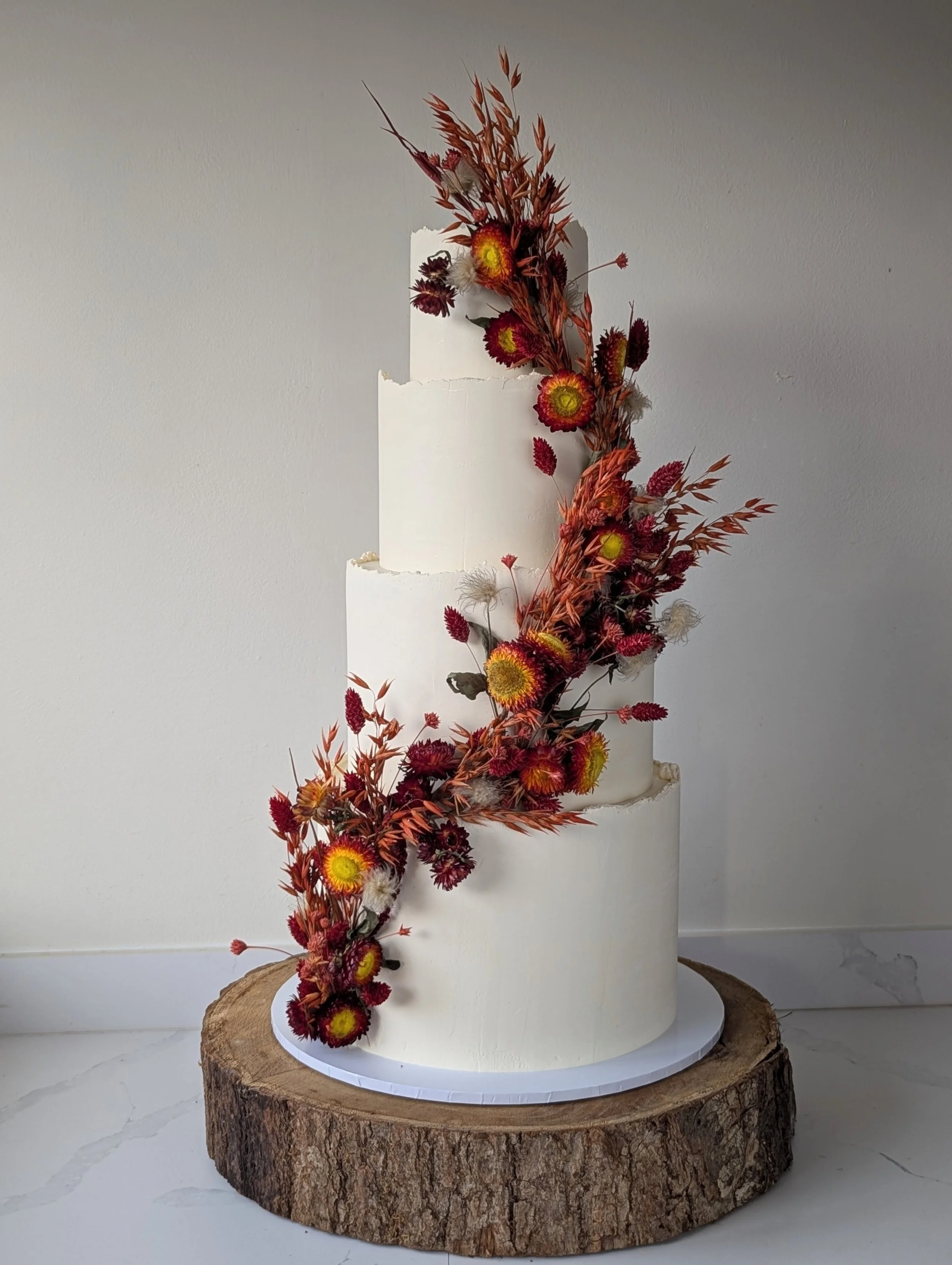 A white tiered cake decorated with dried flowers and grass, placed on a wooden slab. For a beautiful autumnal wedding at Grittleton House. 