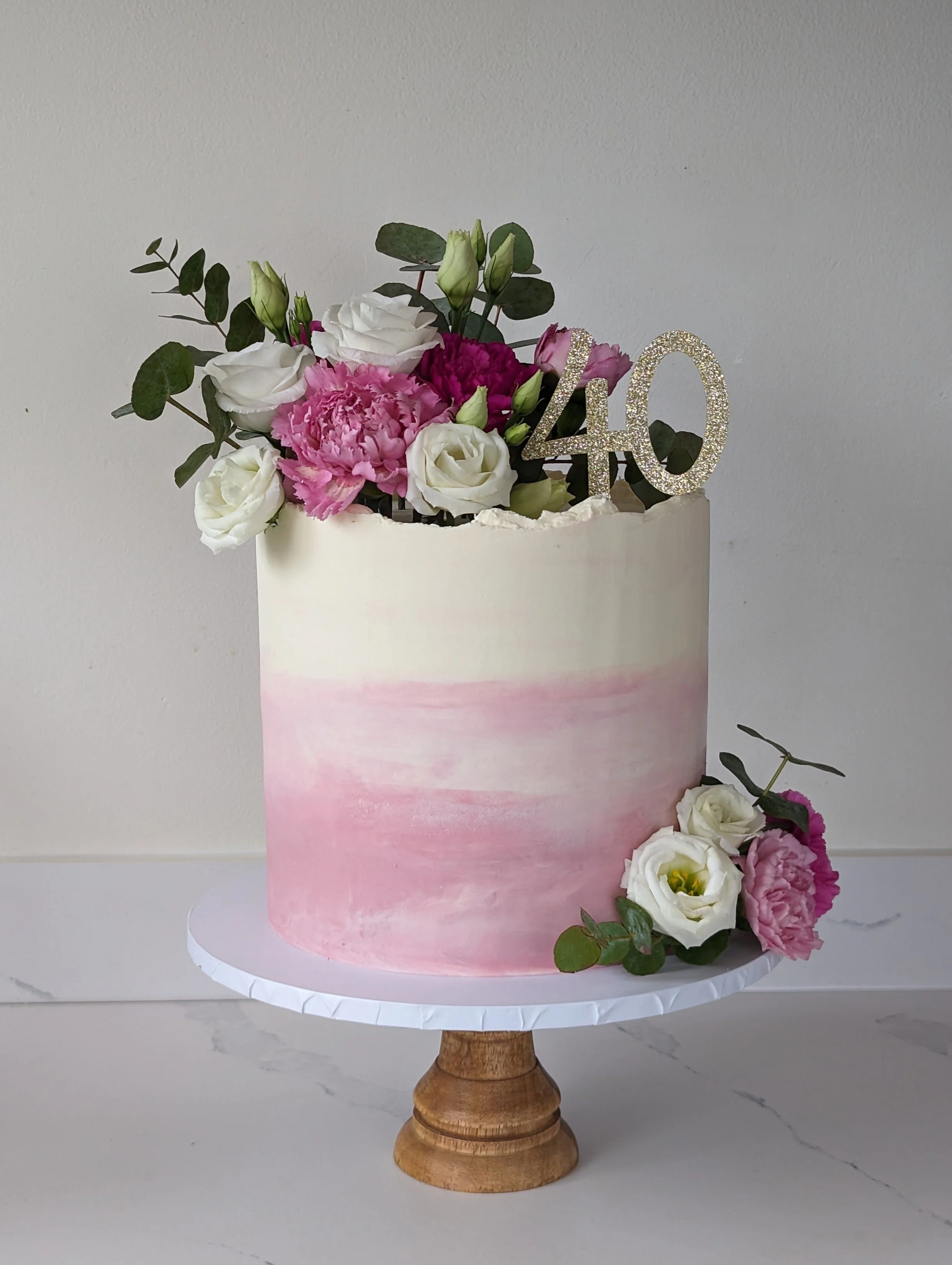 A floral cake decorated with pink and white flowers including roses, peonies, and greenery, with a glittery '40' cake topper, placed on a white cake stand with a wood base.