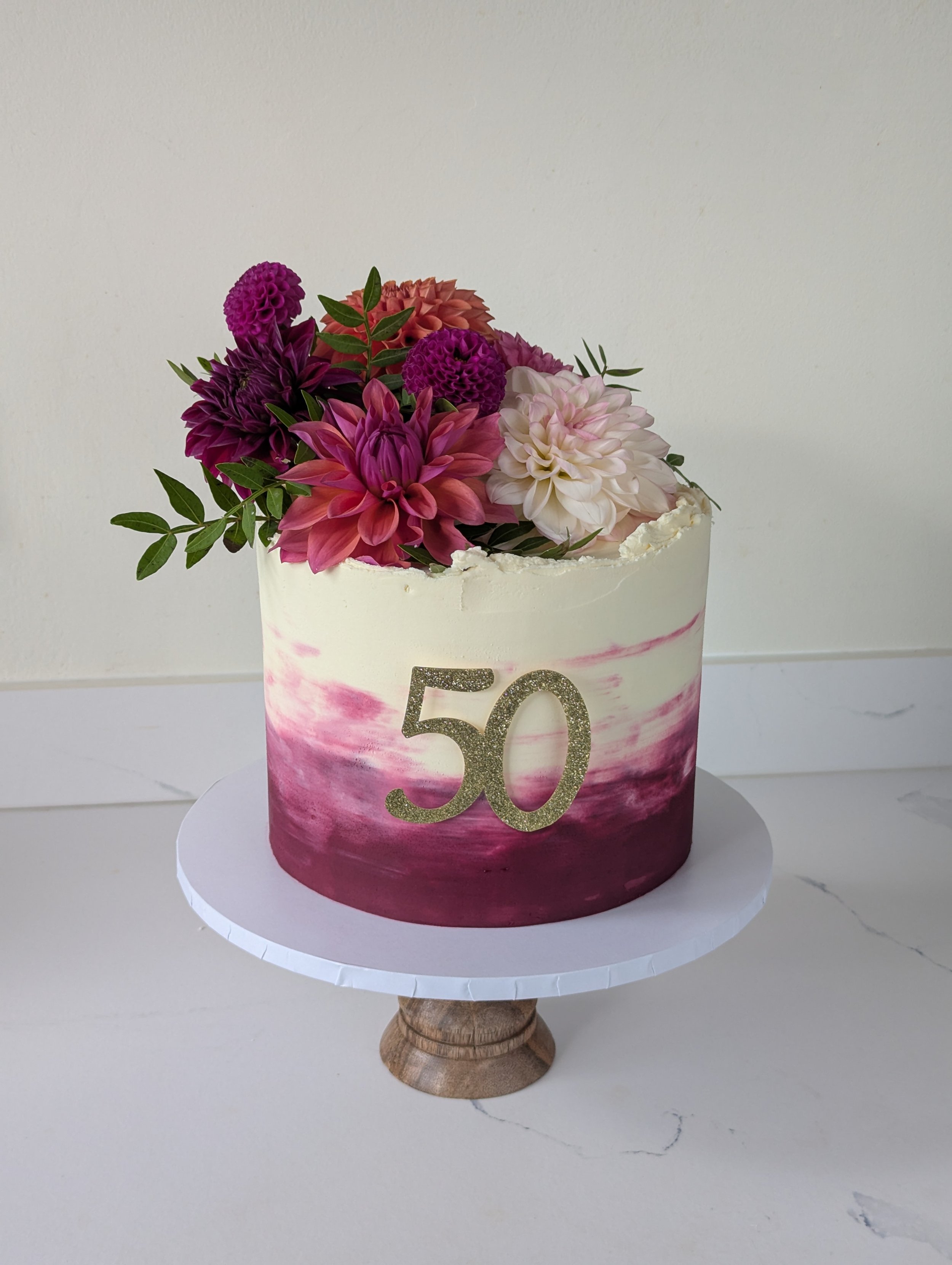 A birthday cake decorated with pink, purple, and white flowers, with the number 50 glittery gold, on a white cake board and cake stand.