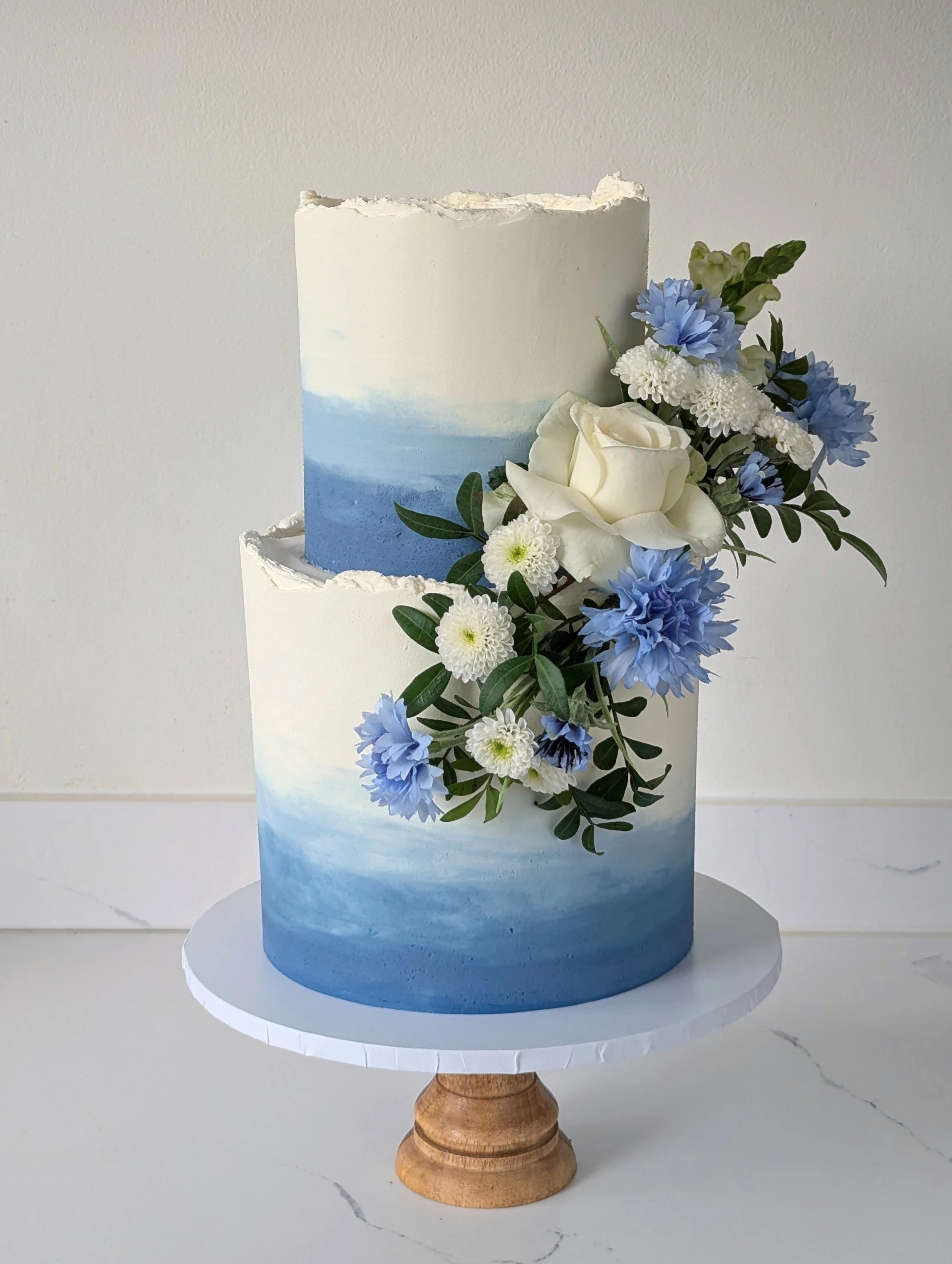 A three-tiered cake with a watercolor ocean scene decorating the middle tier, decorated with a floral arrangement of white roses, blue and white chrysanthemums, and green foliage on the side. Delivered to Bowood wedding. 