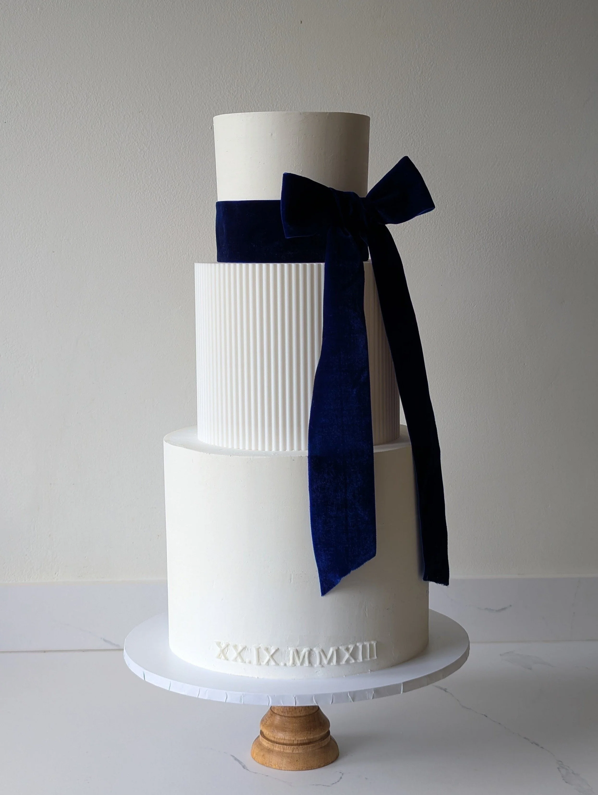 A white tiered cake with textured vertical lines on the middle tier, a dark blue velvet bow on the side, and Roman numerals 'XX X MMXXIII' engraved at the base, placed on a white pedestal against a plain wall.