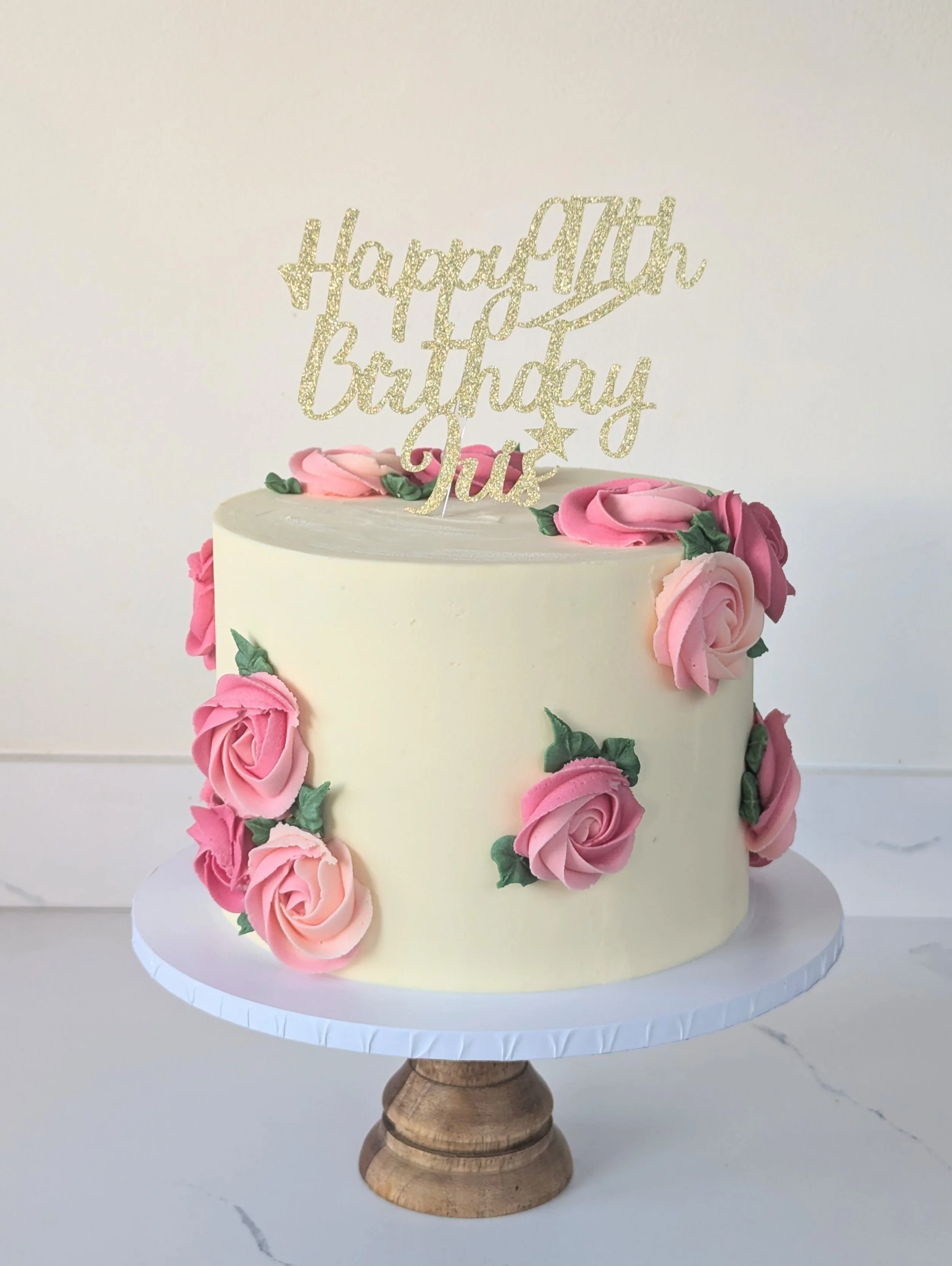 A white birthday cake decorated with pink and white roses and green leaves, with a gold glitter 'Happy 4th Birthday' topper, placed on a white cake stand.