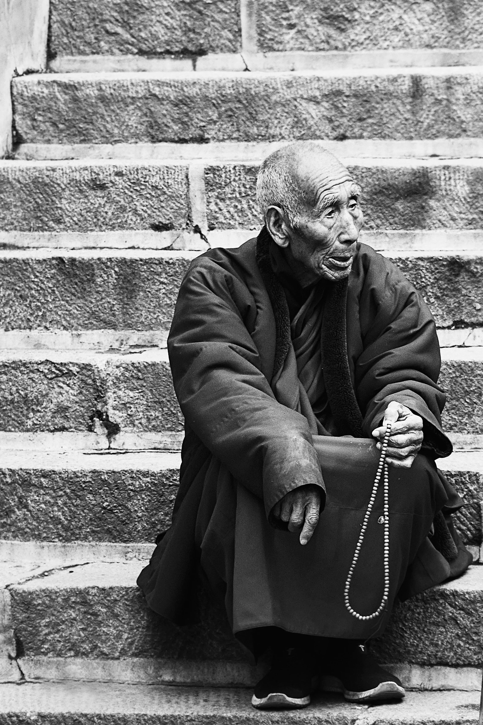 An elderly man with a bald head and wrinkles, wearing traditional robes, sitting on stone steps, holding a rosary or prayer beads in his right hand, with his mouth open.
